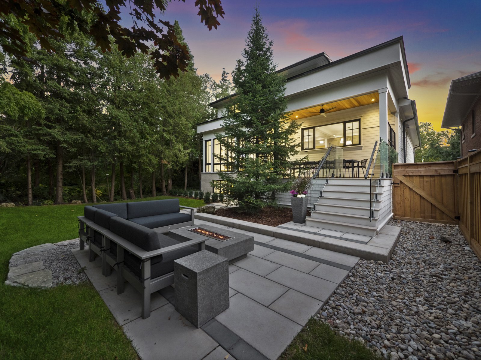 View of a backyard featuring a patio at sunset with a custom built modern house, a black outdoor sofa set around a fire pit, stone pavers, a small staircase, a large evergreen tree, and a wooden fence.