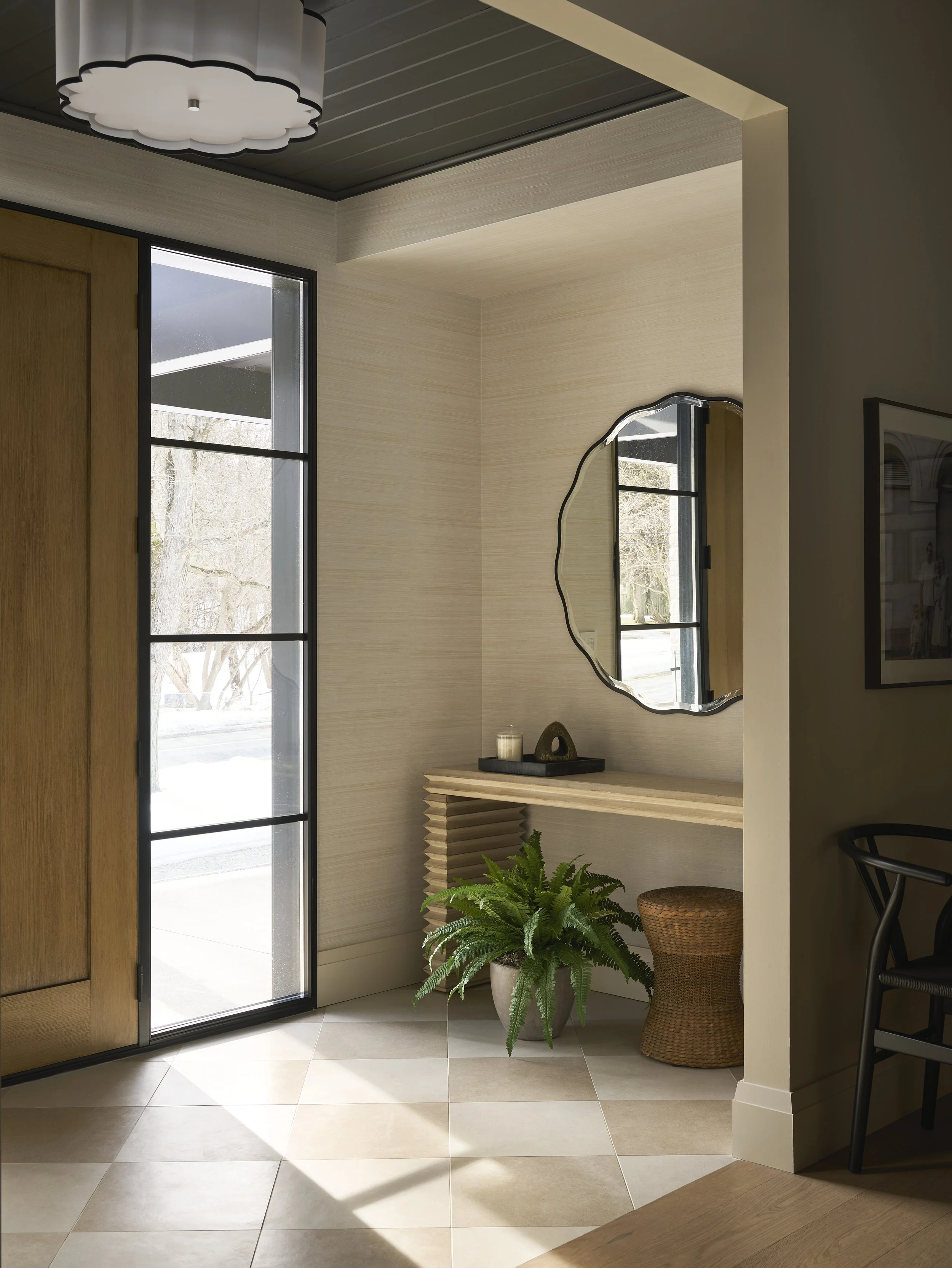 Bright entryway with a large window, a mirror, a console table with a candle and decorative object, a potted fern, and a rattan stool.