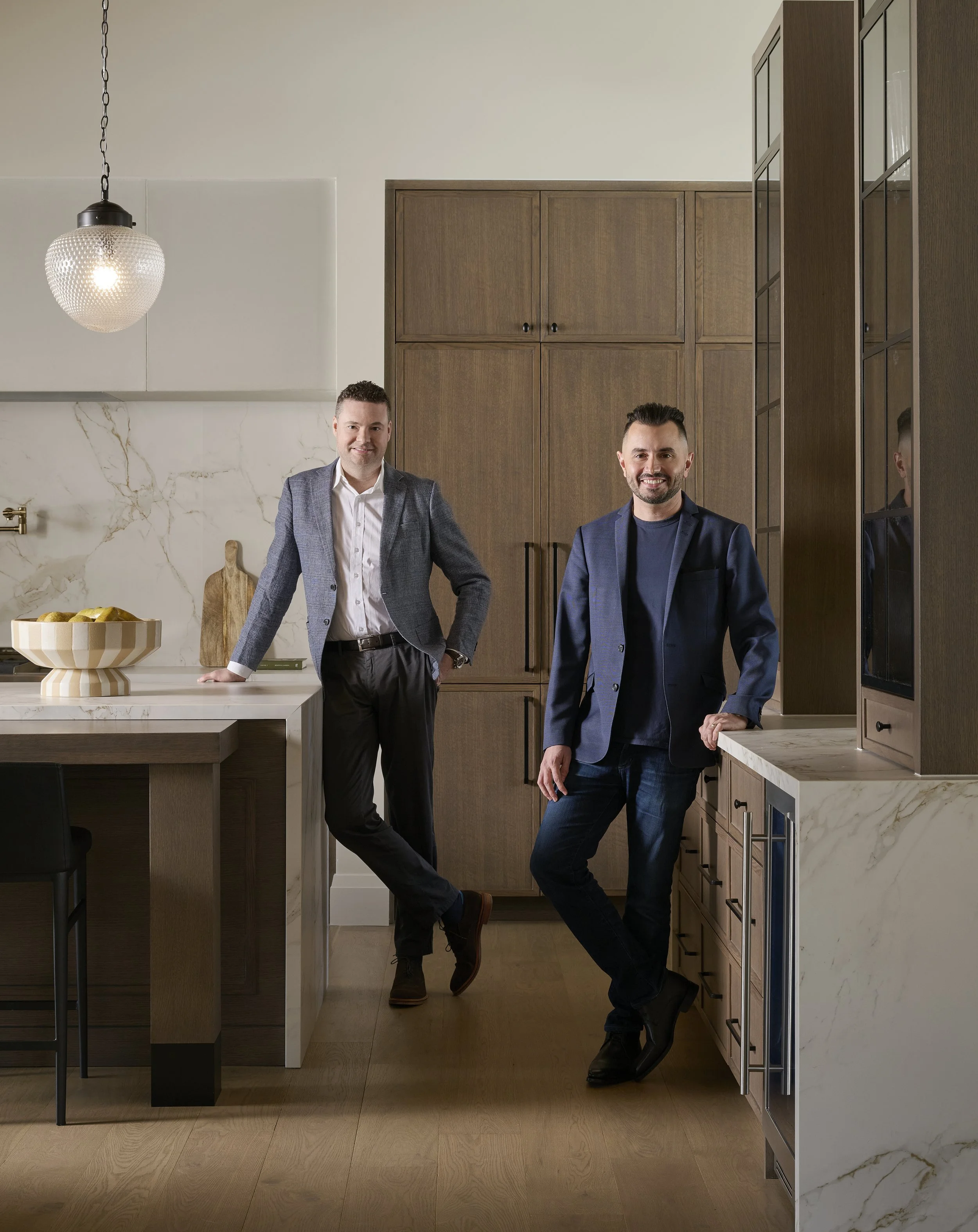 Two men (the founders, Al and Justin)  in suits smile and pose in a modern kitchen with wooden cabinets and marble countertops.