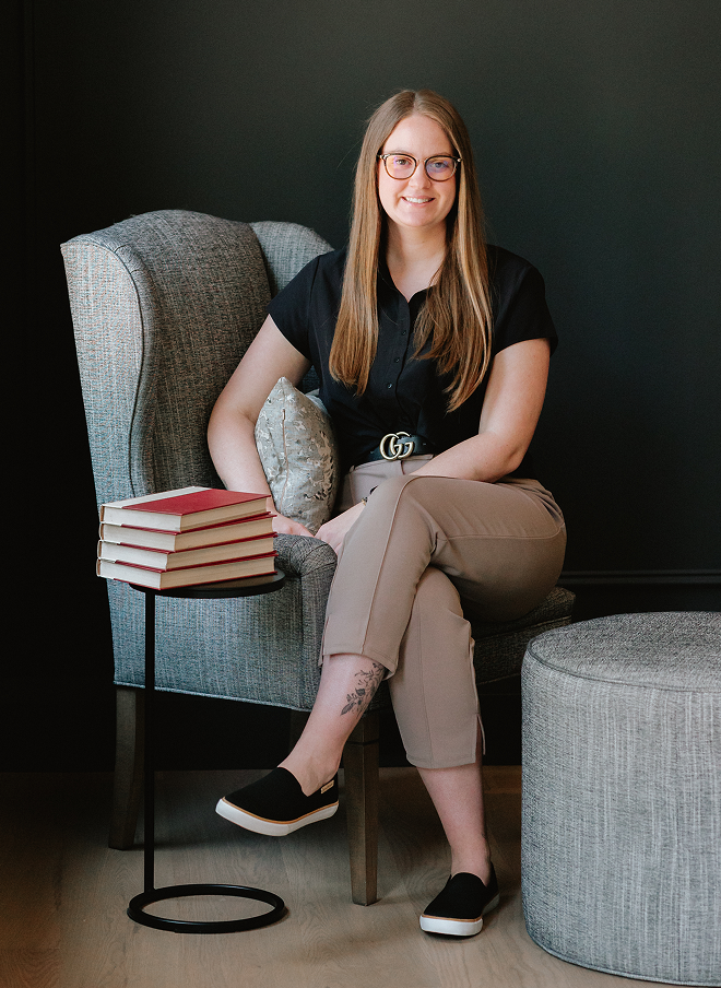 A woman with long hair, glasses, wearing a black shirt, beige pants, and slip-on shoes sitting on a gray armchair with a pillow. She is smiling and has a stack of books on a small round side table next to her. The background is black.