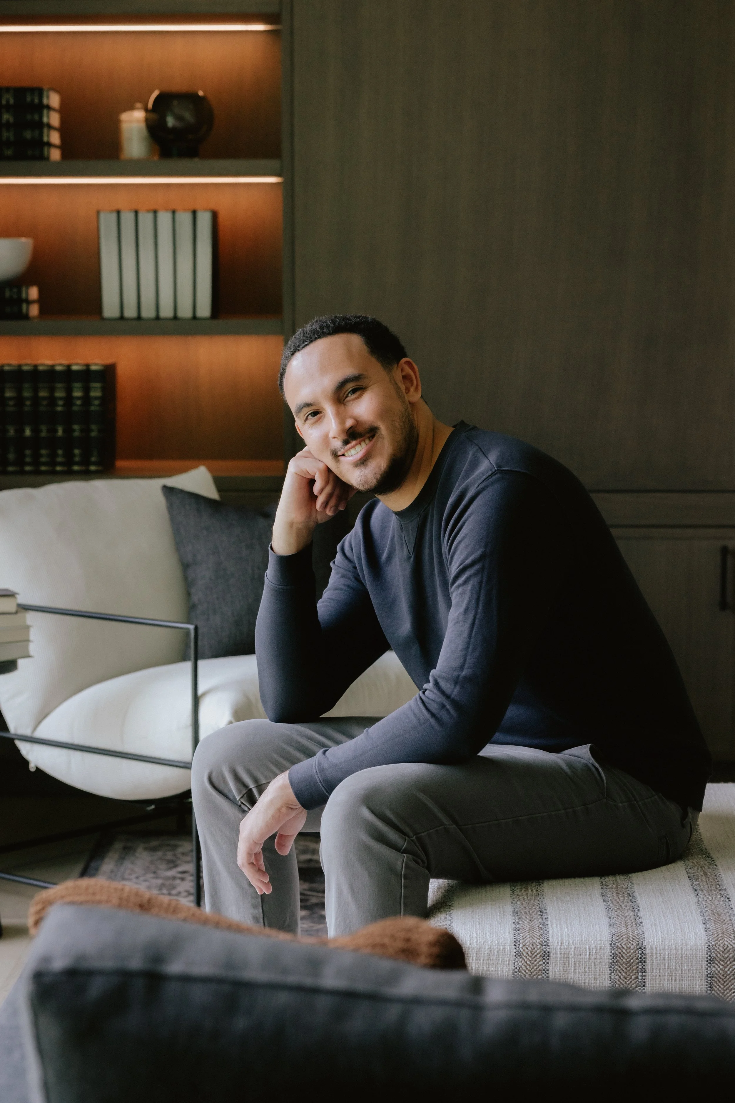 A man with short dark hair and a beard, smiling and sitting on a cushioned bench in a modern, warmly lit living room.