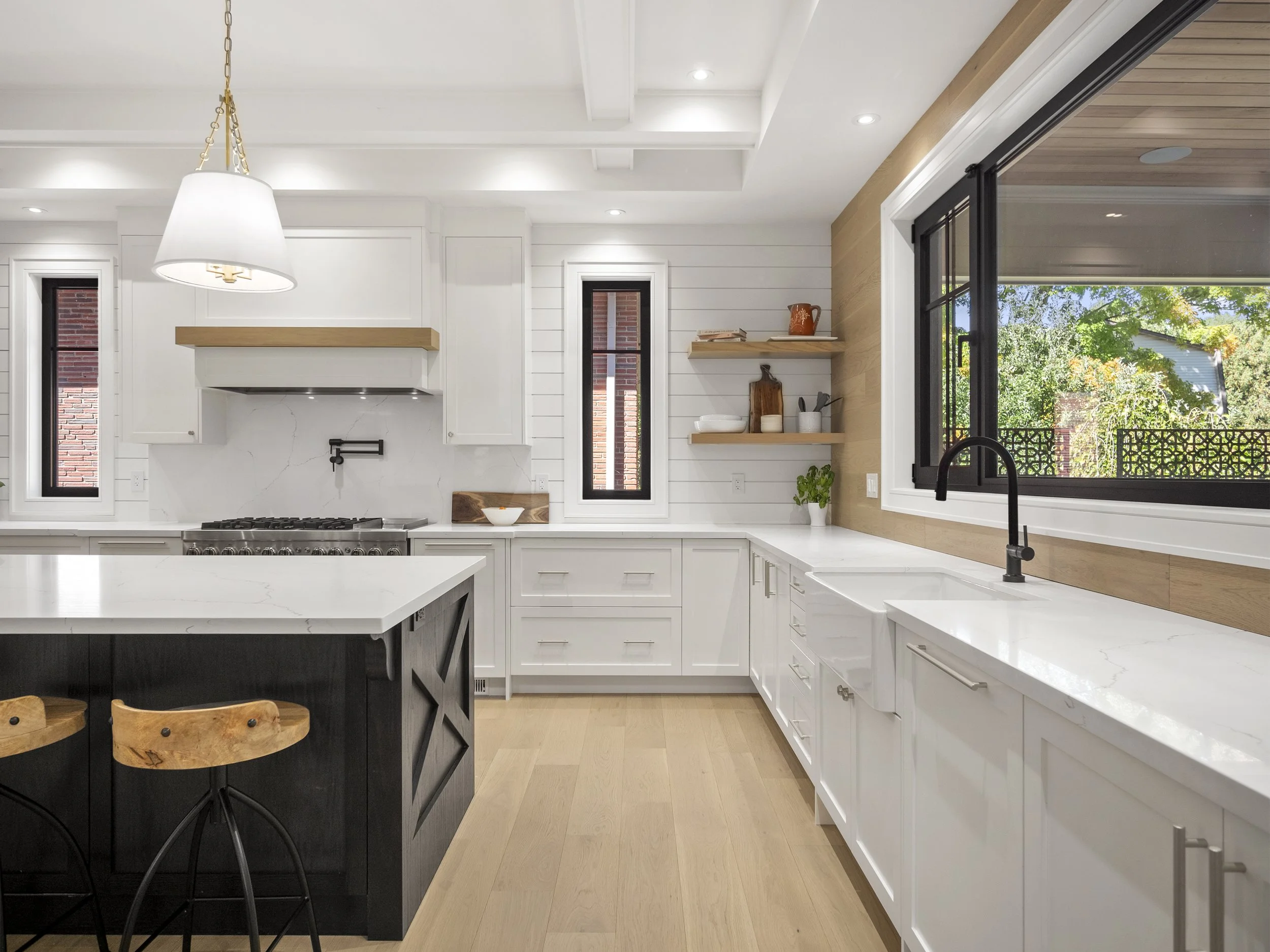Modern custom built kitchen with white cabinets, black faucet, large window showing greenery, and wooden accents.