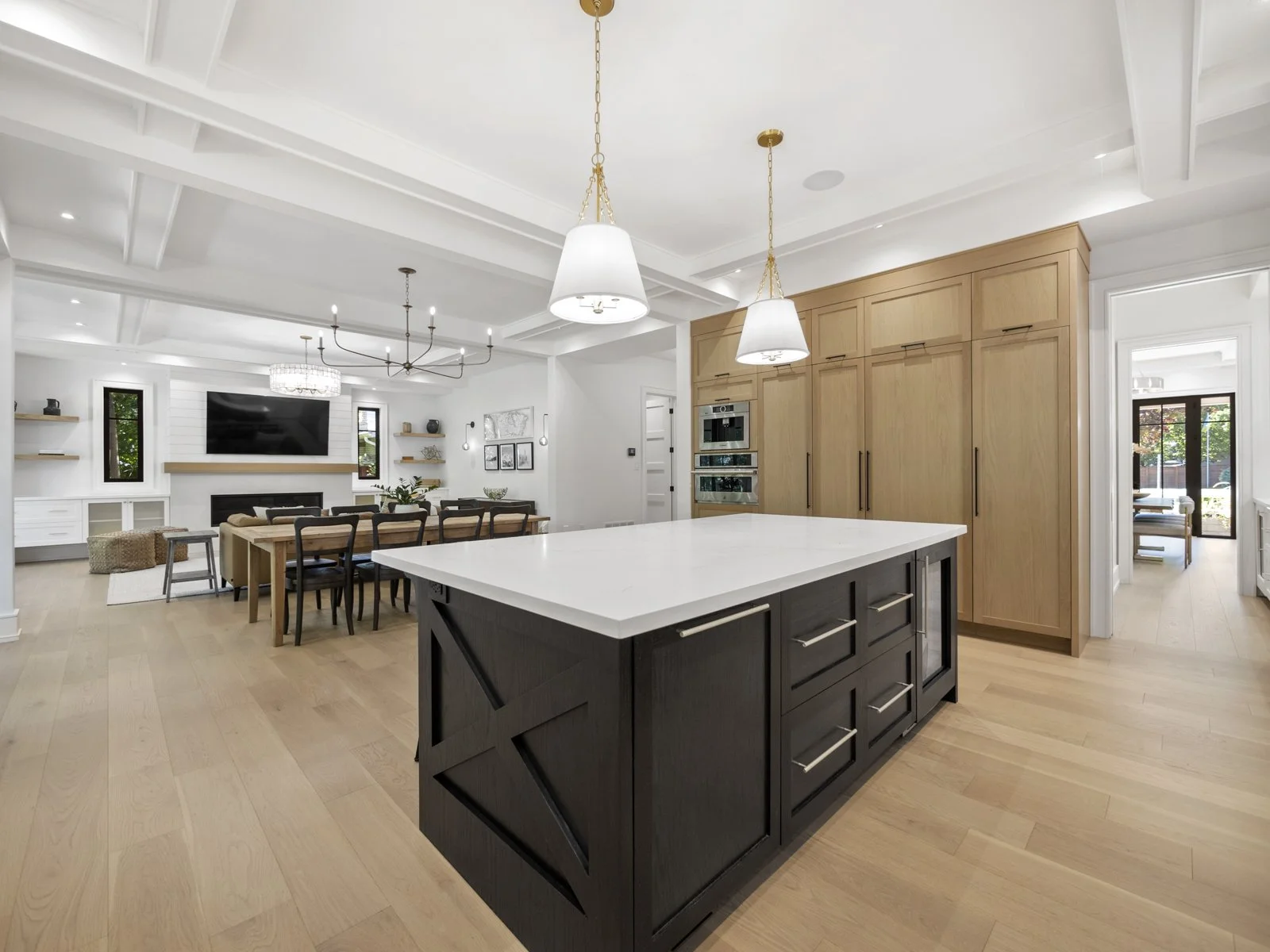 Open-concept kitchen and dining area with light hardwood floors, white walls, and a vaulted ceiling with beams. The kitchen has a black island with a white countertop, pendant lighting, built-in appliances, and wooden cabinetry. The dining table is wood with black chairs, and the living area features a large wall-mounted TV and a chandelier.