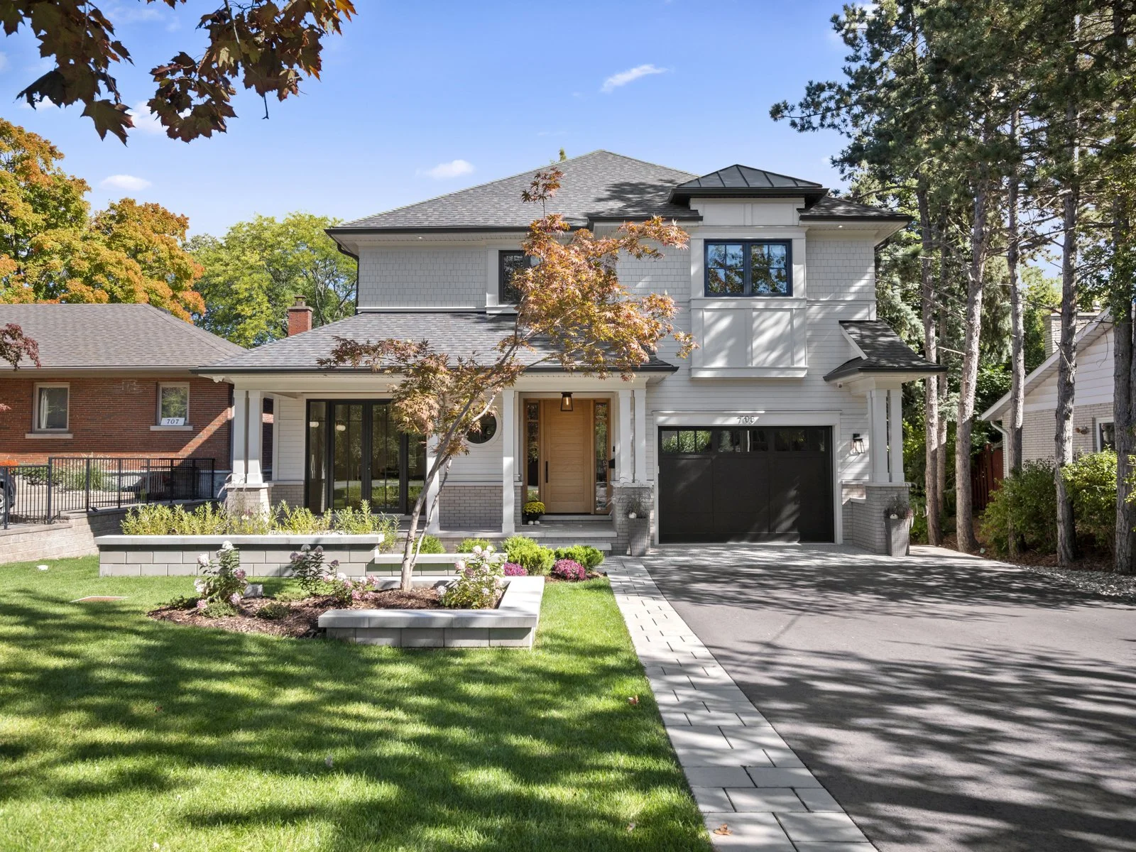 A modern custom built two-story house with white siding, black garage door, and a wooden front door, surrounded by a well-maintained lawn, trees, and bushes under a clear blue sky.