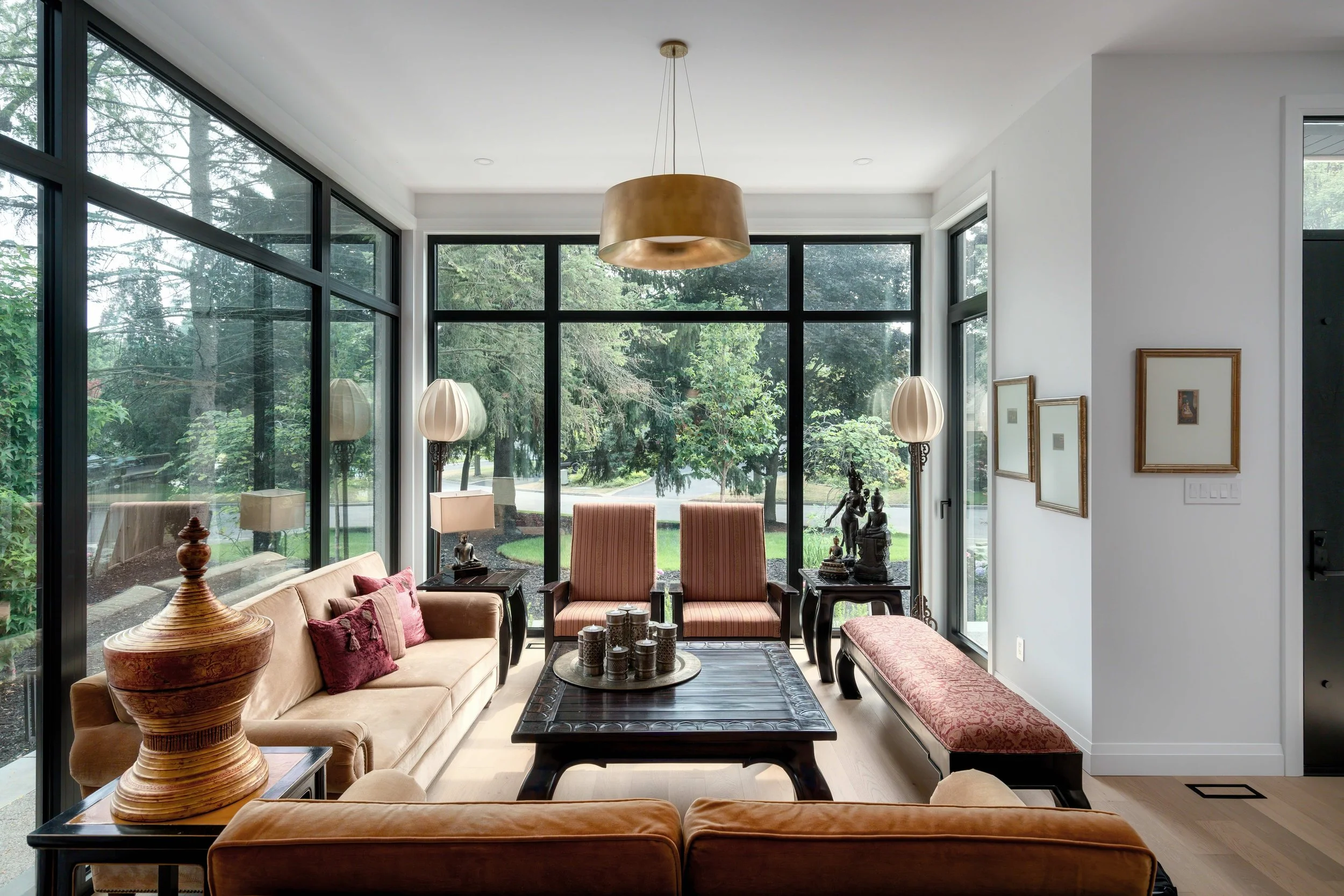 Living room with large windows, beige sofa with red pillows, two pink striped armchairs, a dark wood coffee table with decorative jars, side tables with lamps, and framed artwork on the white walls.