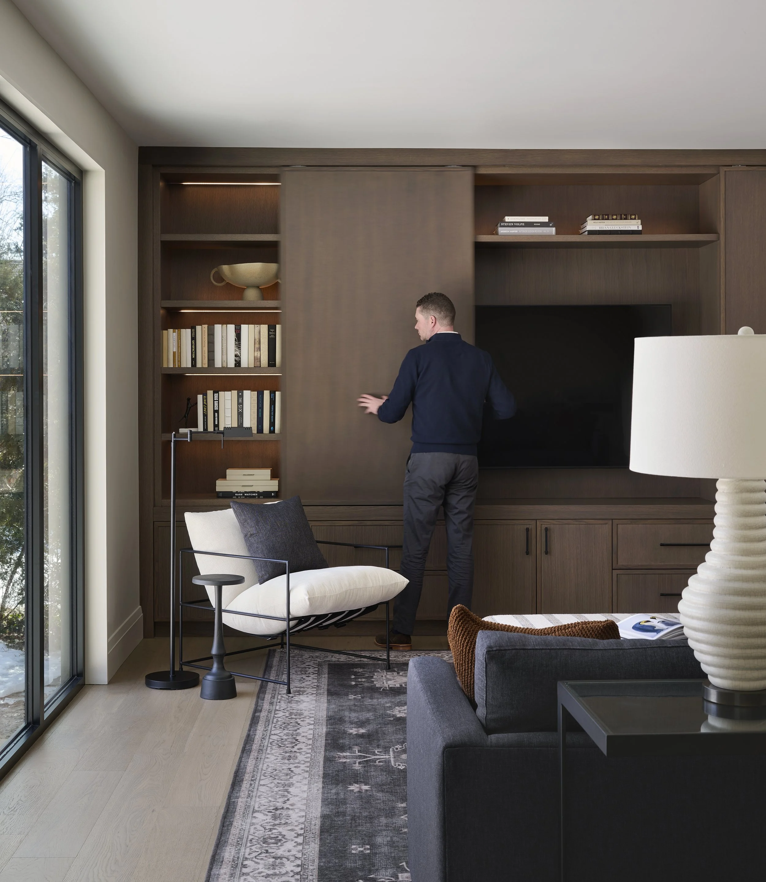 A man in a dark sweater and gray pants standing in a modern living room, looking at a large wooden shelving unit with books and decorative items, near a sliding glass door.