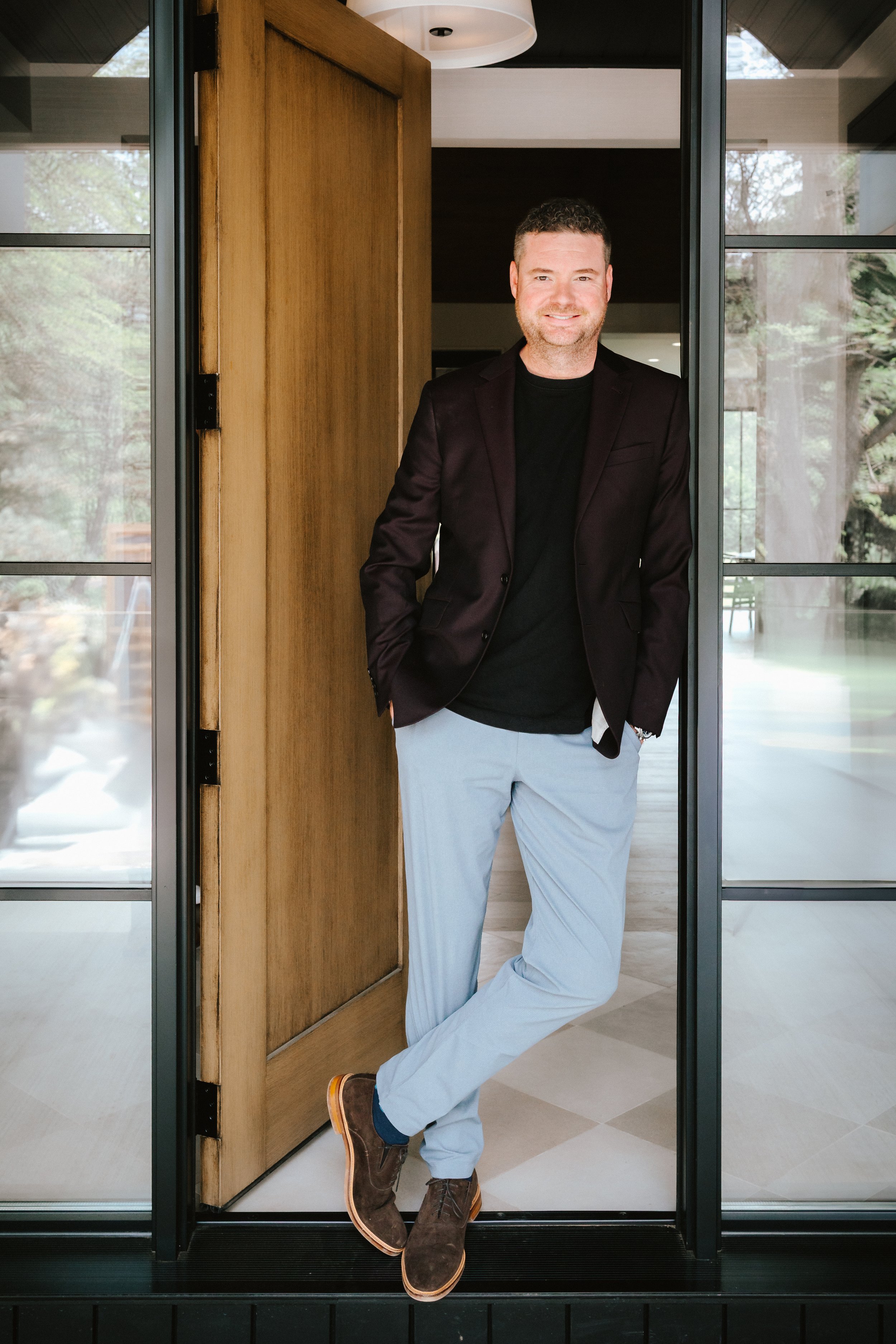 A man with short dark hair, beard, wearing a black shirt, purple blazer, light gray pants, and brown shoes, standing in a doorway with a smile.