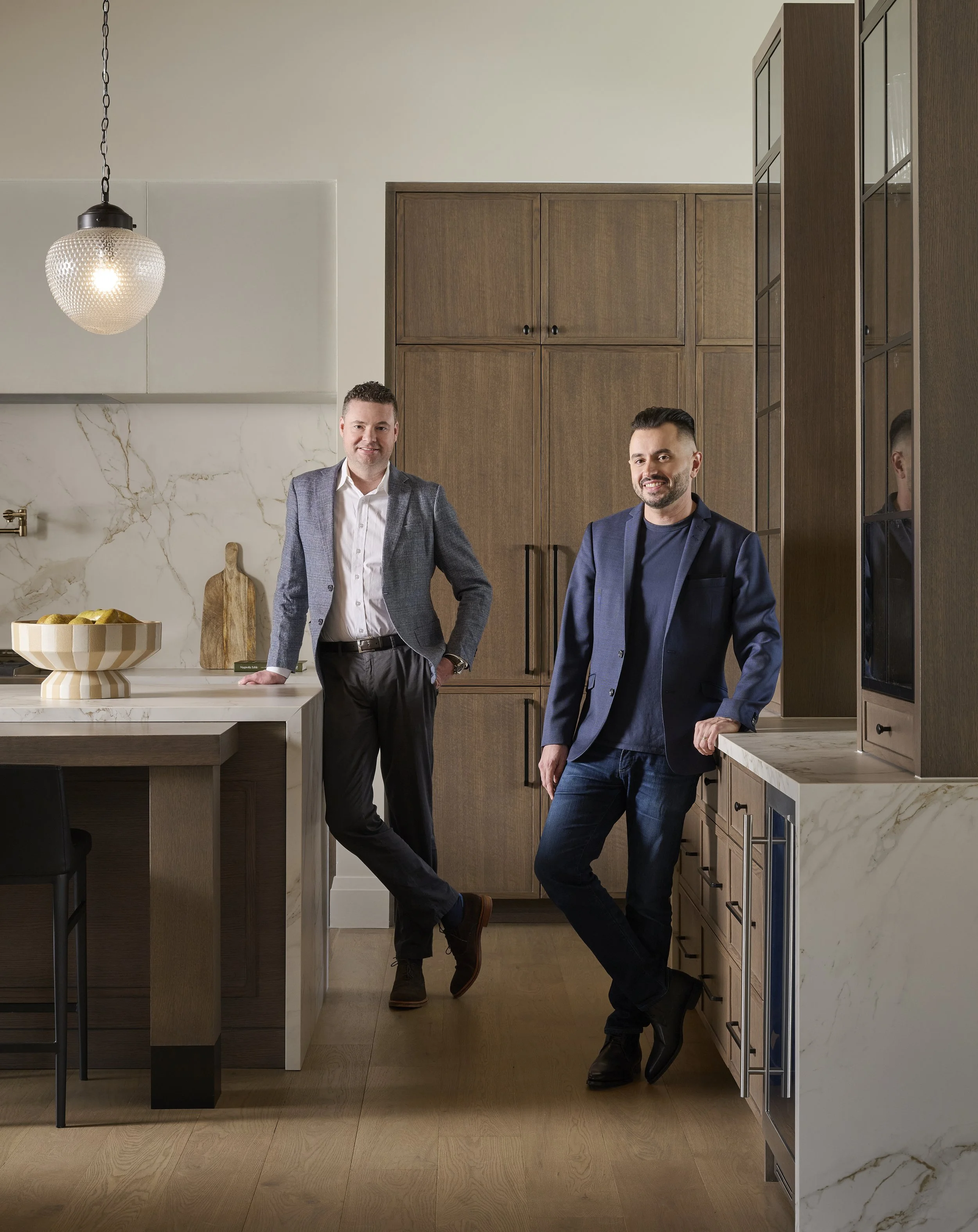 Two men (the founders, Al and Justin) in business casual attire standing in a modern kitchen, leaning against a kitchen island and cabinets.