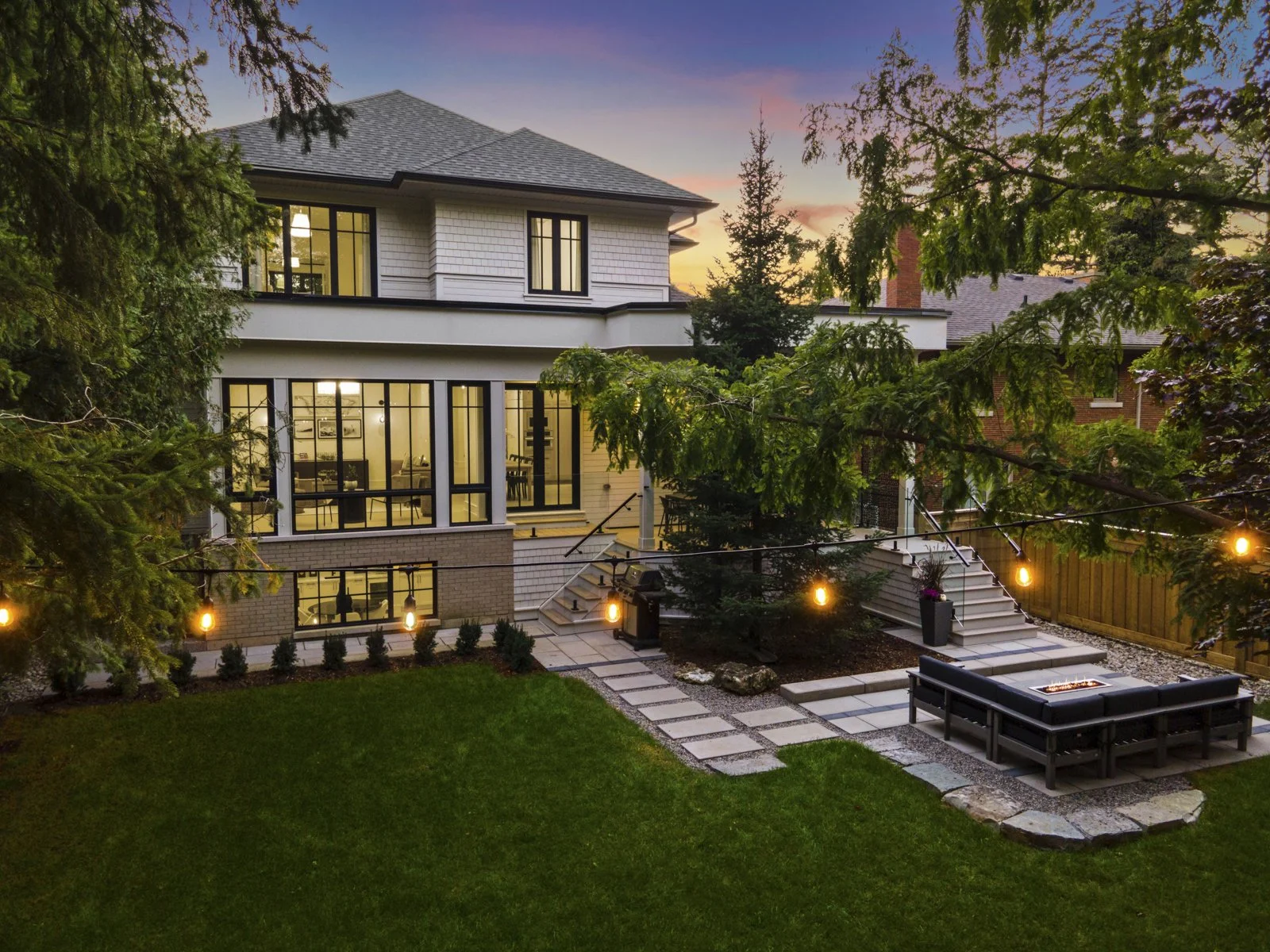A modern two-story custom built house at dusk, with large windows lit from inside, surrounded by trees and a well-maintained yard with a stone path, outdoor seating area with a fire pit, string lights, and a staircase leading to the upper level.