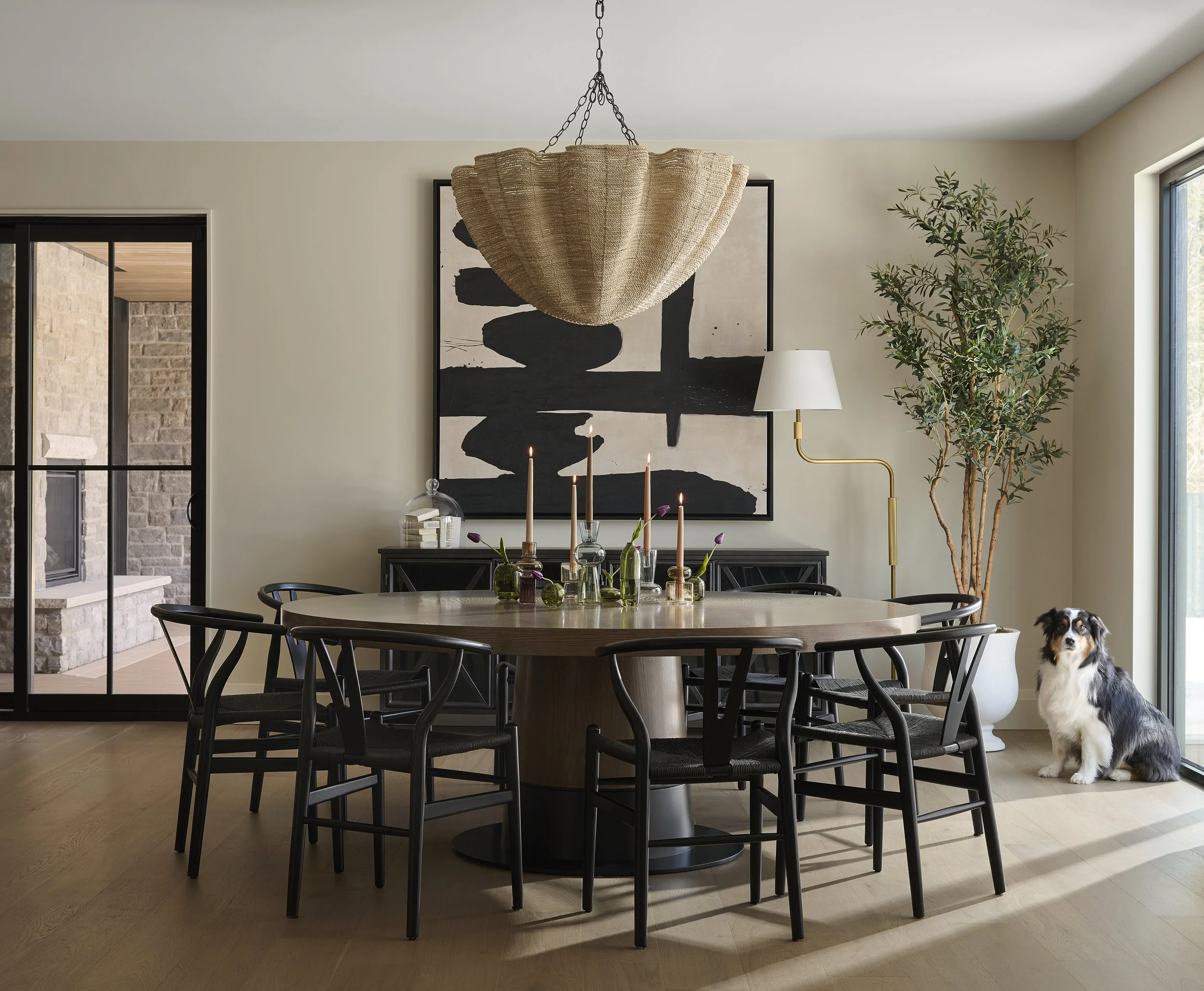 Modern dining room with a round table, black chairs, candle holders, a large black and white abstract painting, a tall floor lamp, and a large potted plant. A very cute dog is sitting near the sliding glass door.