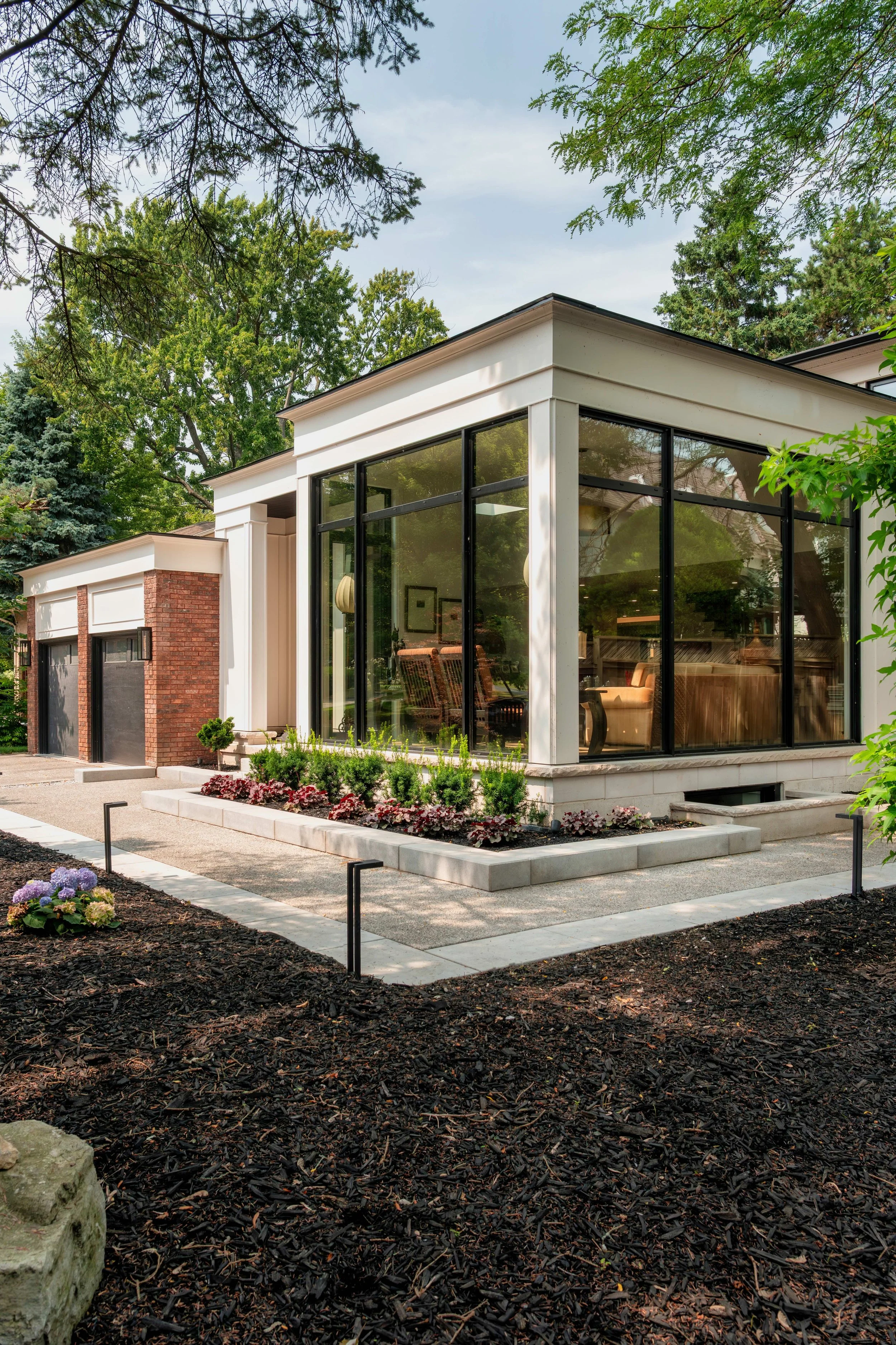 Modern house with custom built addition featuring large glass windows, brick and white siding exterior, landscaped front yard with flower bed and mulch, and a driveway with garage doors.