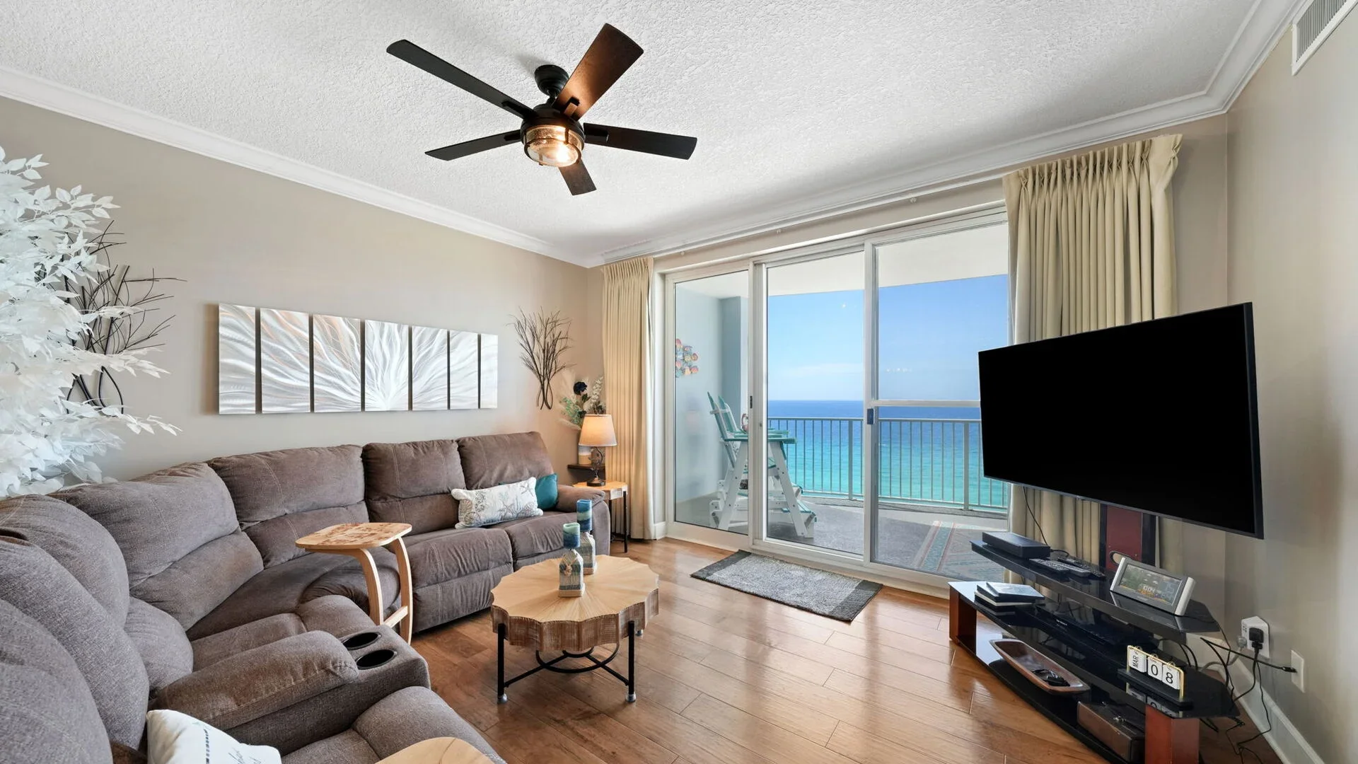 Living room with large sliding glass door leading to balcony and overlooking ocean, includes sofa, side table with lamp, wall art, and flat-screen TV.