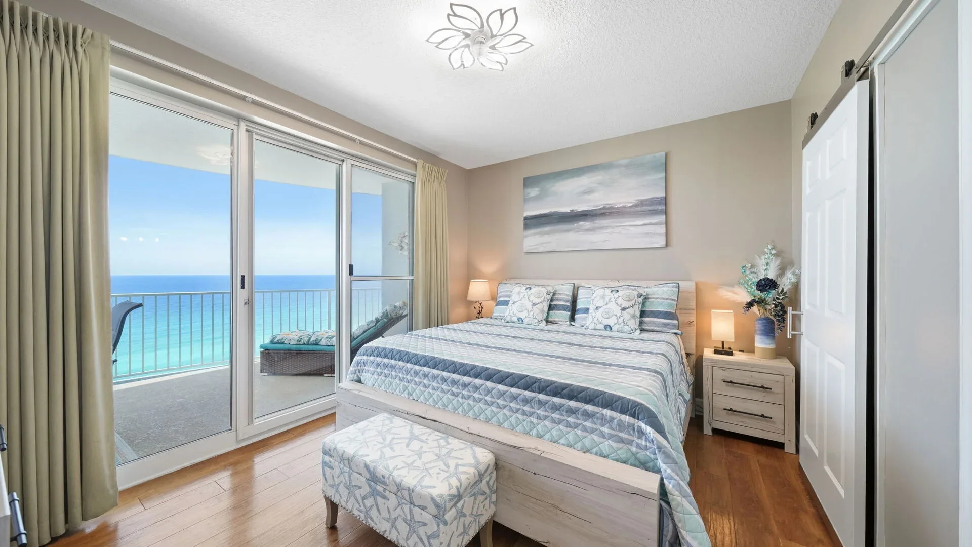 A bedroom with sliding glass doors leading to a balcony overlooking the ocean. The bed has blue and white bedding and pillows, with a small bedside table holding a lamp and a vase with flowers. There is artwork above the bed and an ottoman with a starfish pattern at the foot of the bed. There are cream-colored curtains on the door, and the flooring is wooden.