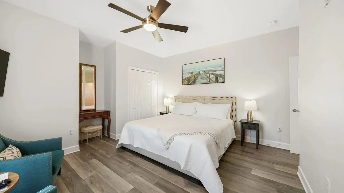 Bright bedroom with king-sized bed, white bedding, two nightstands with lamps, a ceiling fan, and wall art above the bed.