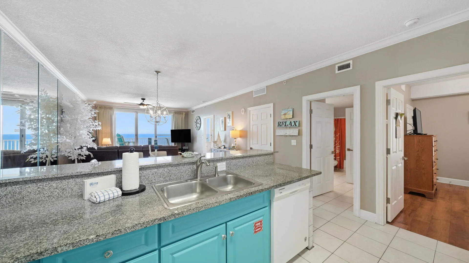 Open-concept living space with a kitchen, visible in the foreground, featuring turquoise cabinets and a granite countertop. Beyond are the living room with a dark couch and large windows showing the ocean view. The interior is decorated with coastal-themed art and signs, with a whirl of color and light from the windows.