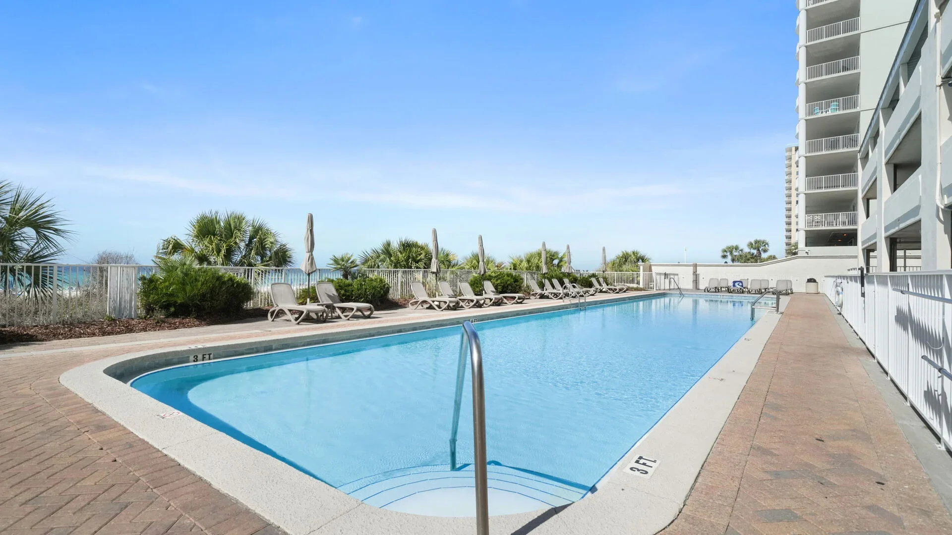 Empty outdoor swimming pool with clear blue water, surrounded by lounge chairs, umbrellas, and greenery, with a tall building in the background under a blue sky.