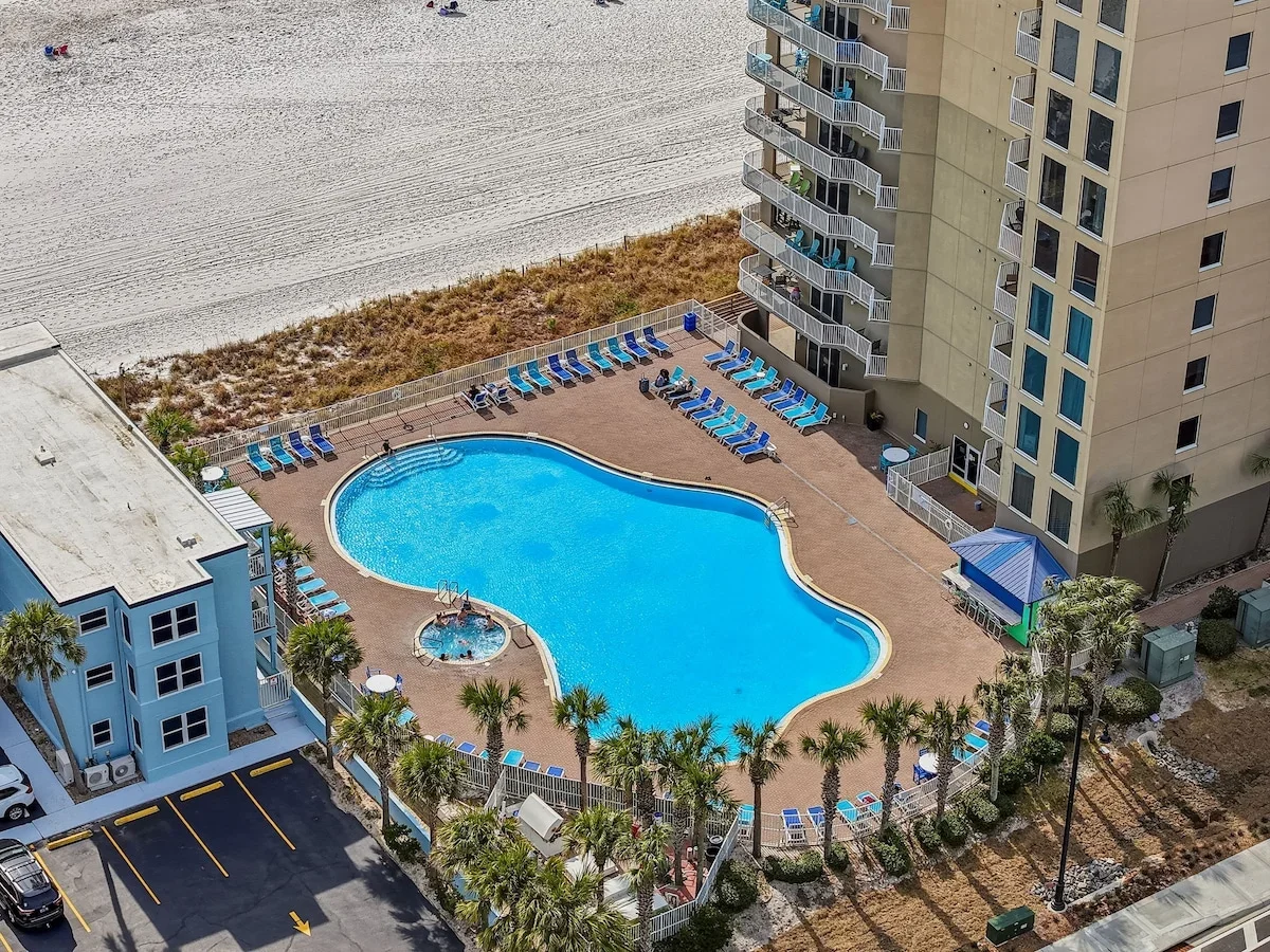 High-angle view of an outdoor swimming pool surrounded by lounge chairs, a small hot tub, an apartment building with balconies, and a beach with sand and beachgoers in the background.