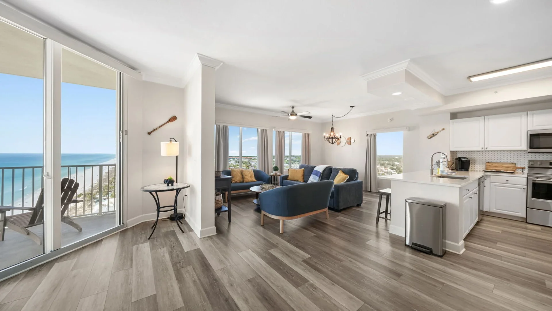 Living room with ocean view, blue sofas, wooden coffee table, kitchen with white cabinets, stainless steel appliances, and a balcony with chairs and ocean scenery in the background.