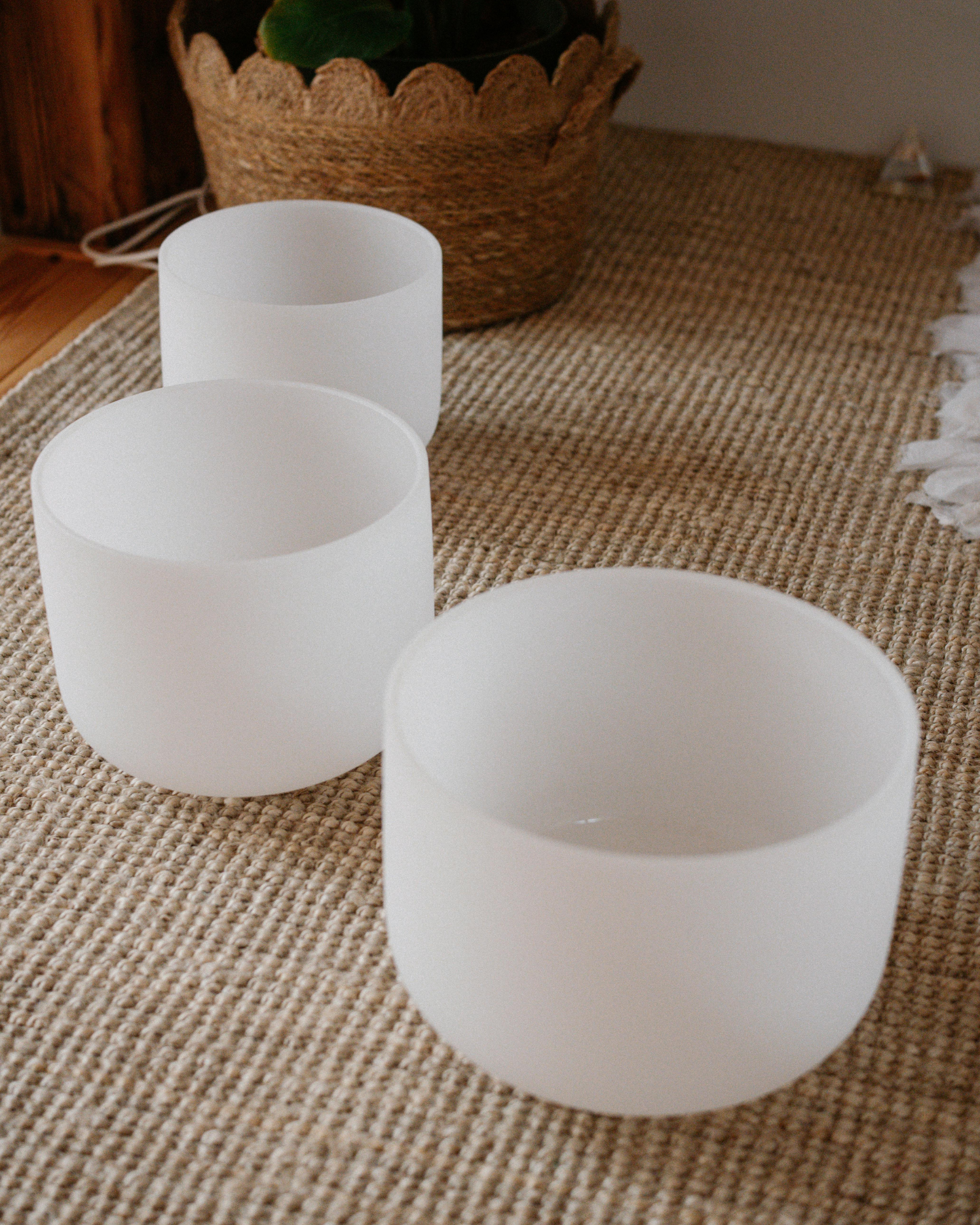 Three white cylindrical candle holders of different heights on a woven beige placemat, with a potted plant in a textured basket in the background.