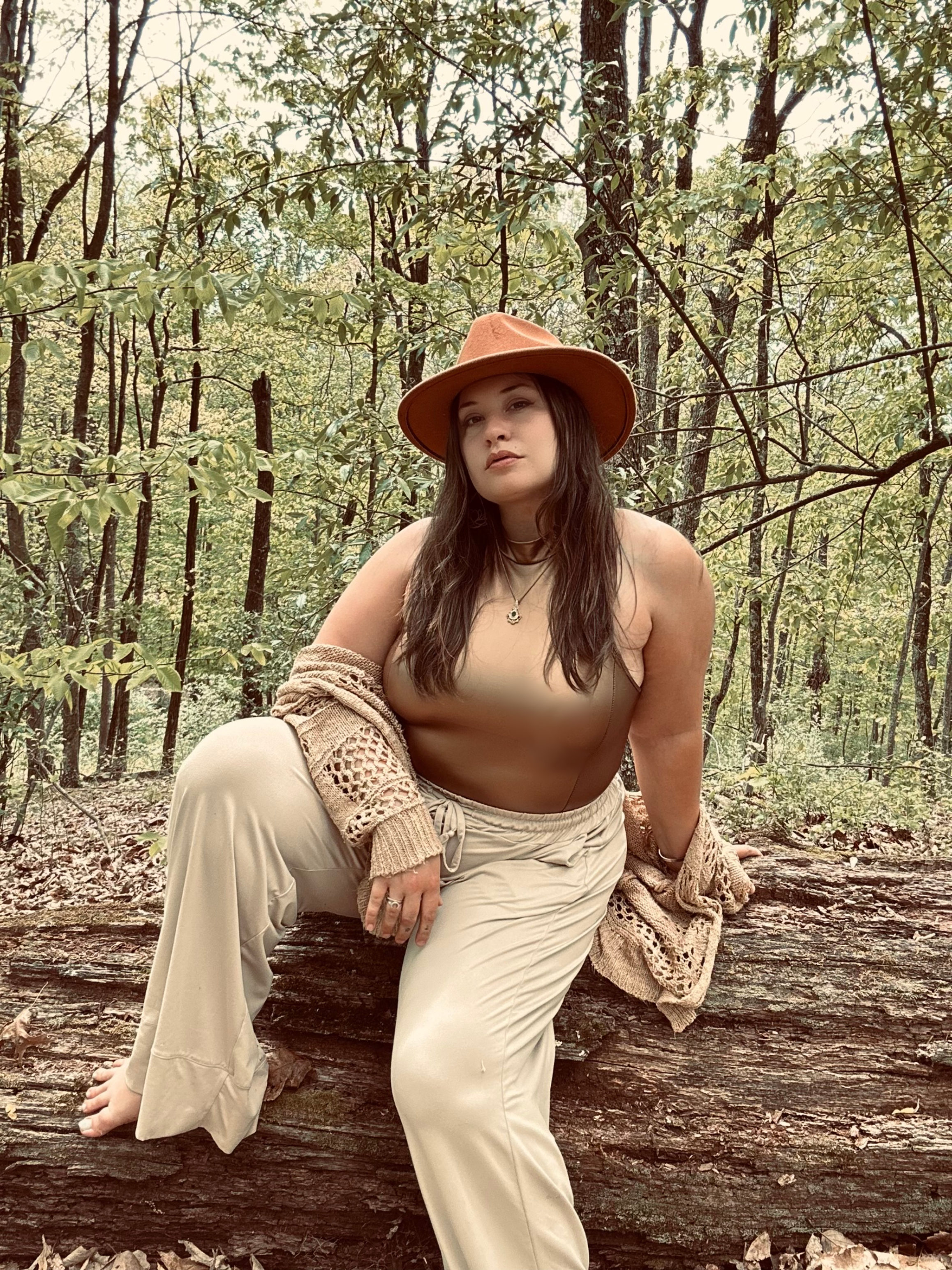 A woman with long dark hair, wearing a beige hat and beige clothing, sitting on a fallen tree trunk in a wooded area with trees and green leaves in the background.