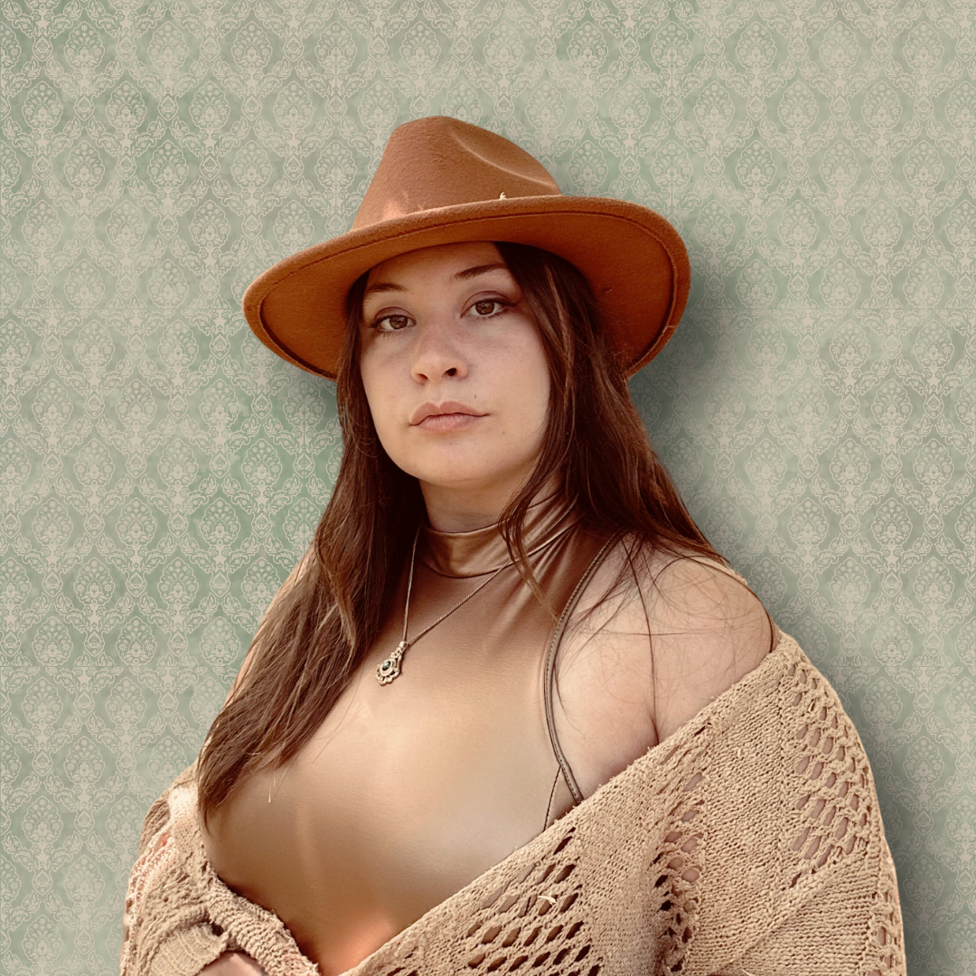 A woman with long brown hair wearing a brown hat, a gold sleeveless top, and a beige crochet shawl, standing against a textured, light green patterned wall.