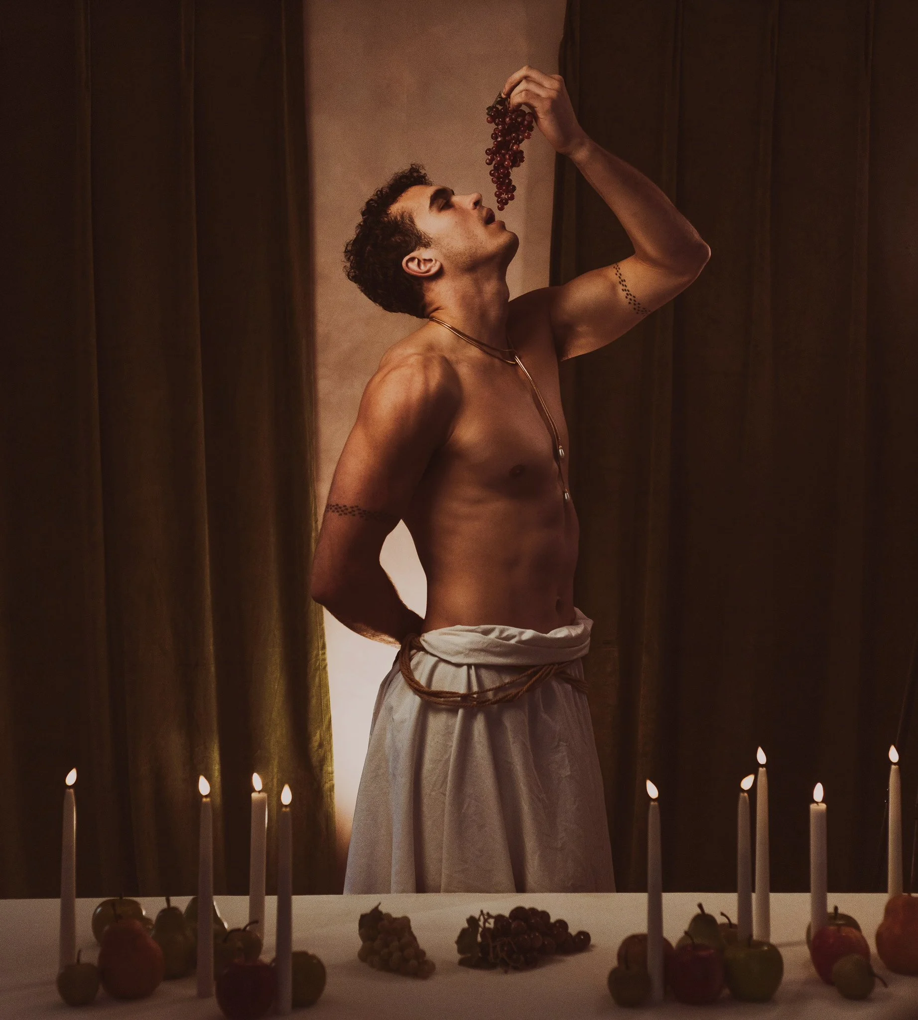 A shirtless man with curly hair and tattoos on his arms stands behind a table with apples, grapes, and lit candles. He is holding a bunch of grapes above his mouth, preparing to eat them in a dimly lit room with dark curtains.
