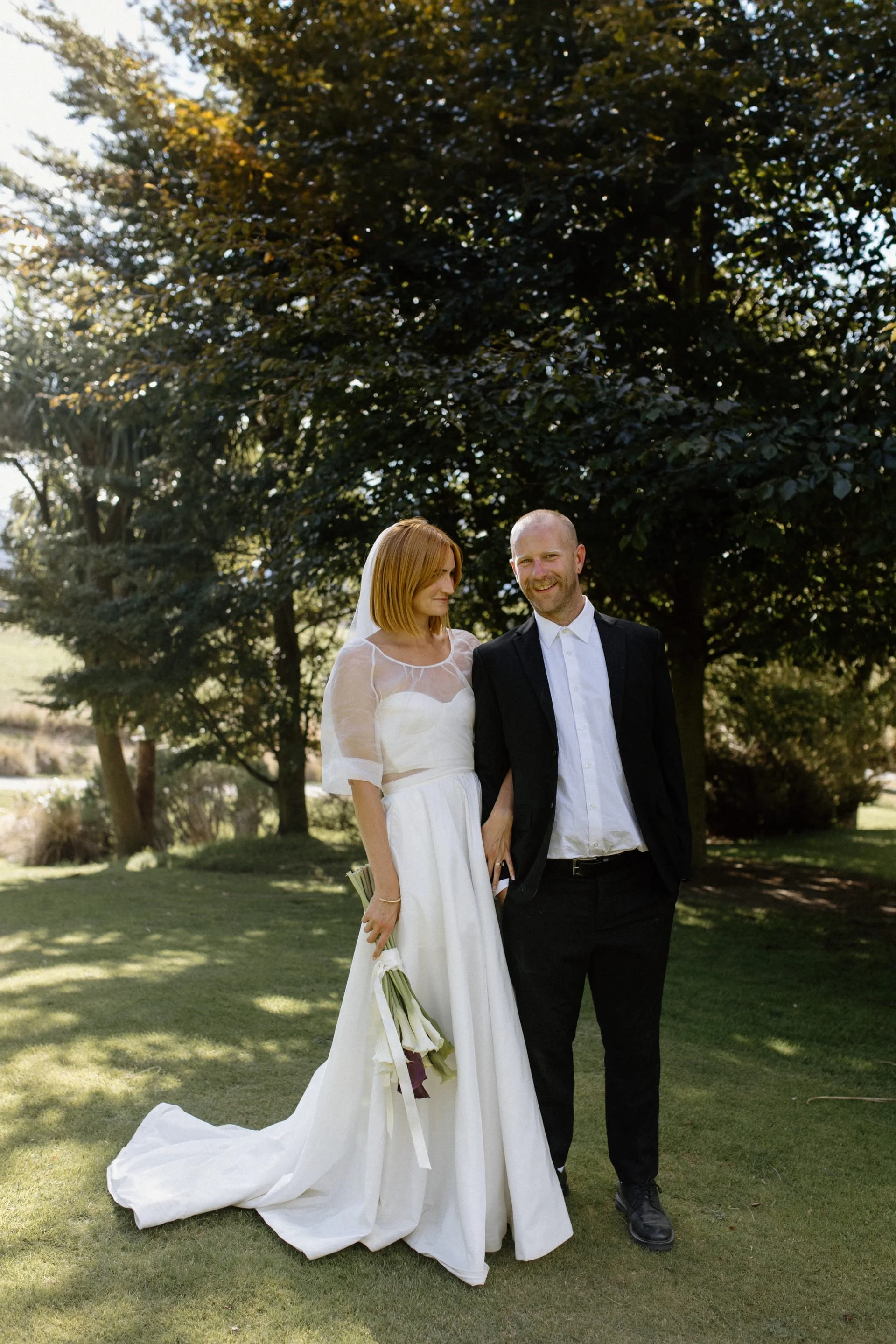 A bride and groom share a glance in the shade of a tree on a summers wedding day in Wanaka