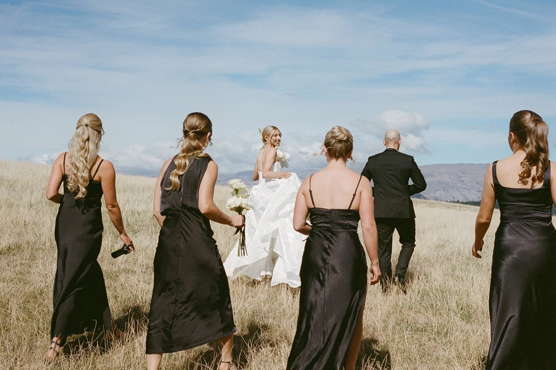 a 35mm film photo of a bride and groom and their bridal party walking through long golden grass at their wanaka wedding held at dublin bay