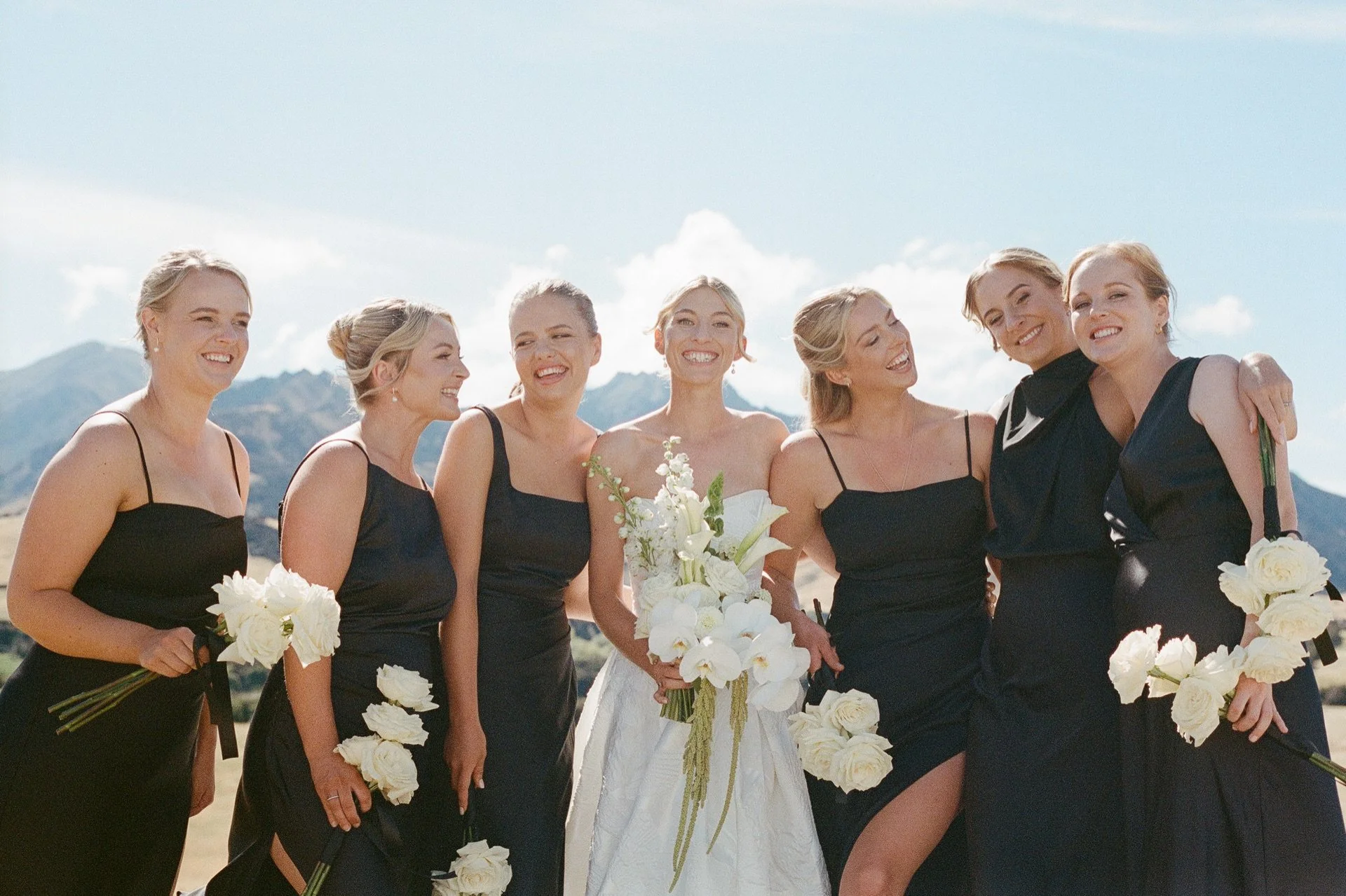 A bride and her bridesmaids cuddle in close with the mountains behind them on a summers wedding at dublin bay in wanaka