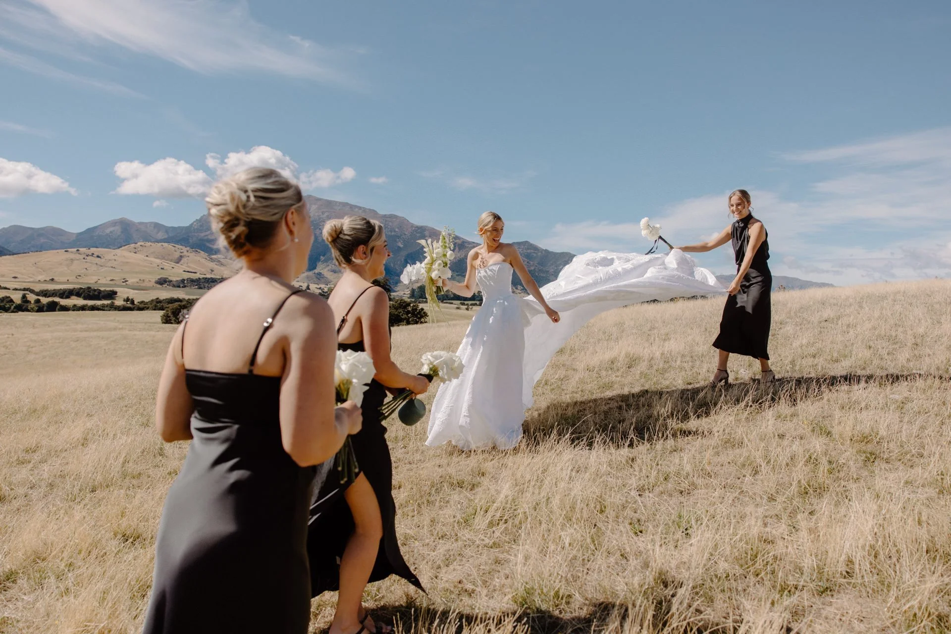 A bridesmaid flicks the brides white dress high in the sky in a field with long golden grass in wanaka at a summers wedding at dublin bay