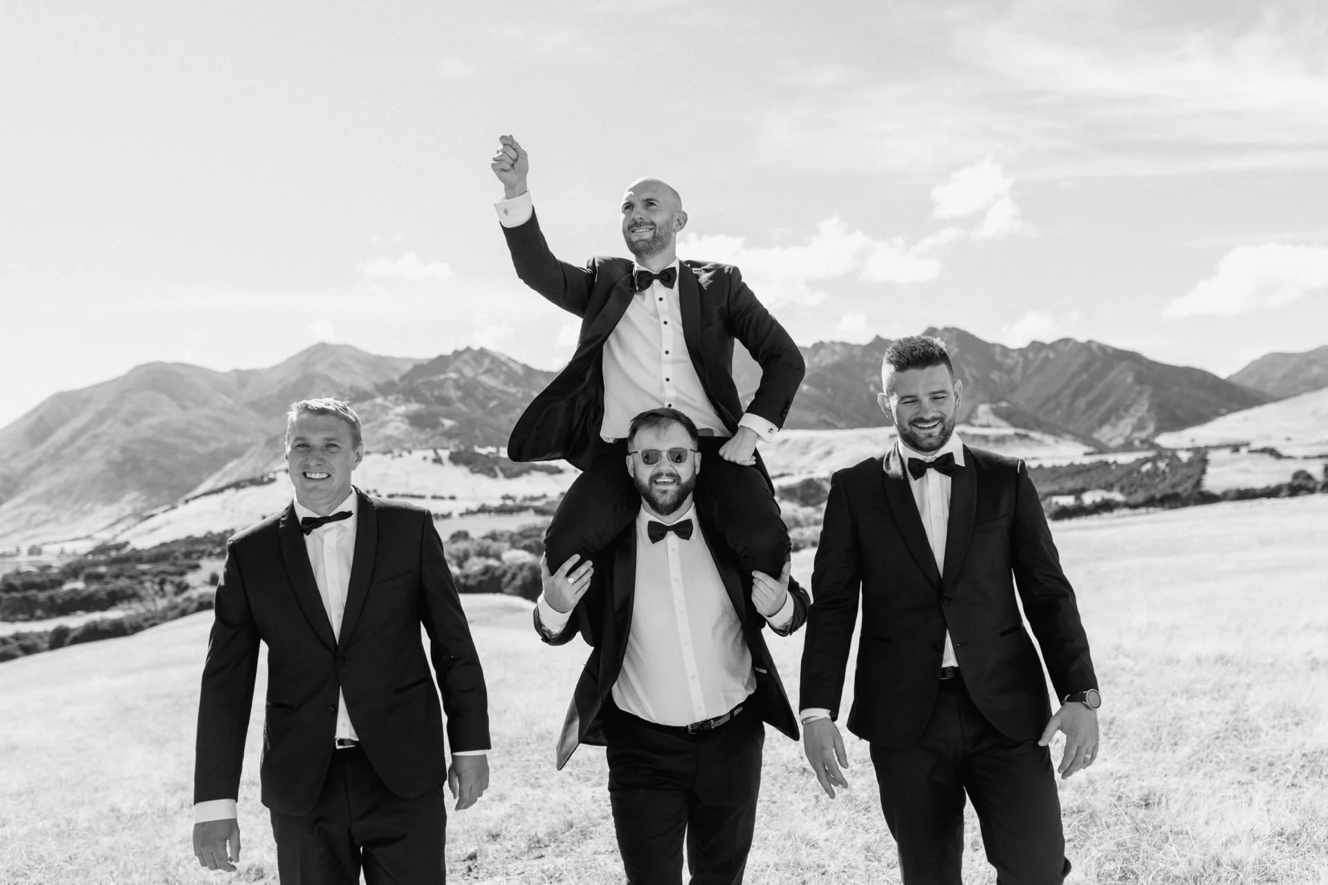 A black and white photo of a groom on the shoulders of his groomsmen walking through a field with mountains towering behind them on their wanaka wedding day.