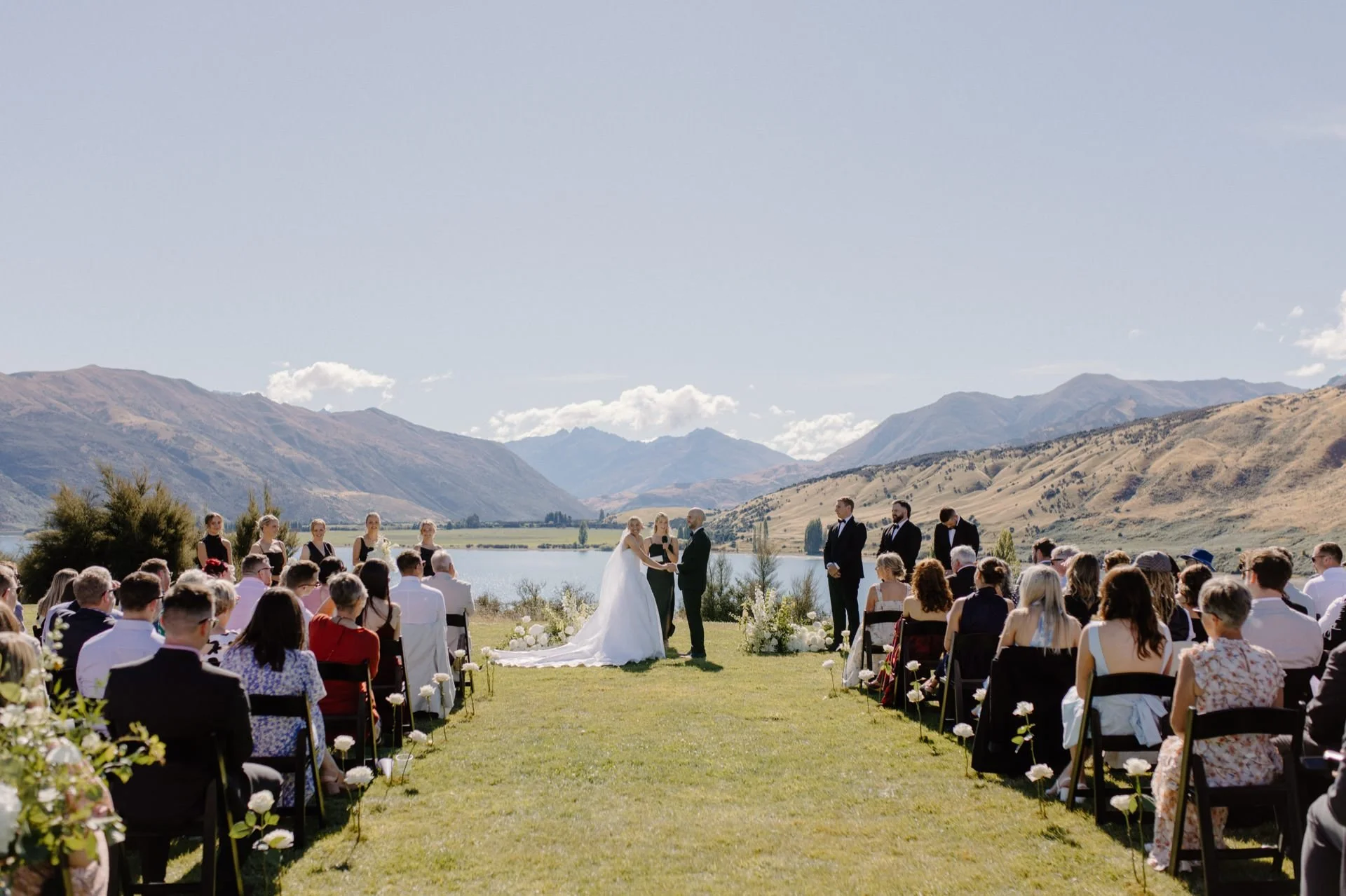 A bride and groom smile at their guests standing at their ceremony held at dublin bay in wanaka at their summer wedding