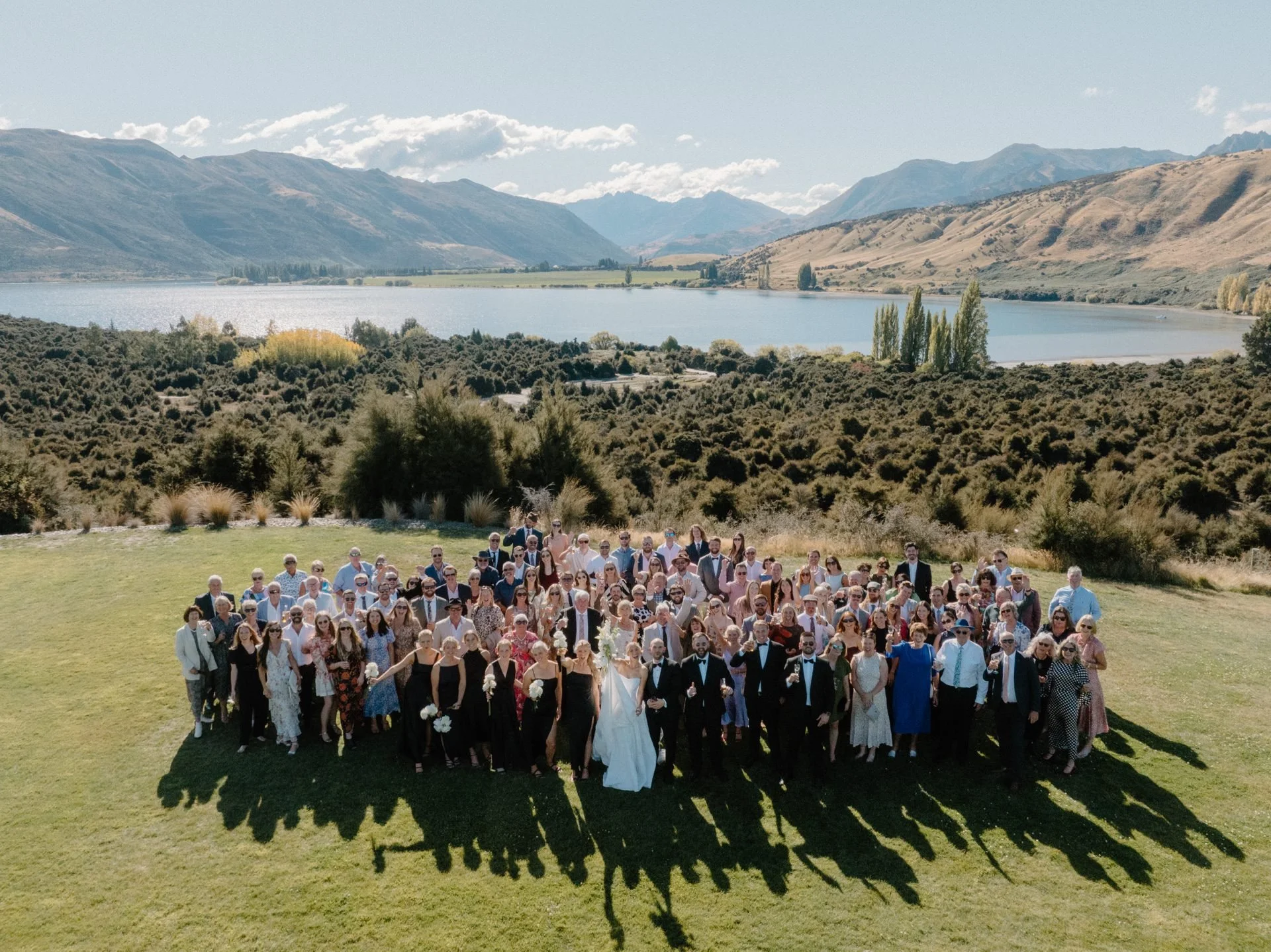 An aerial photo of all guests with the bride and groom showing lake wanaka and the mountains behind them at dublin bay on their summer wedding day.
