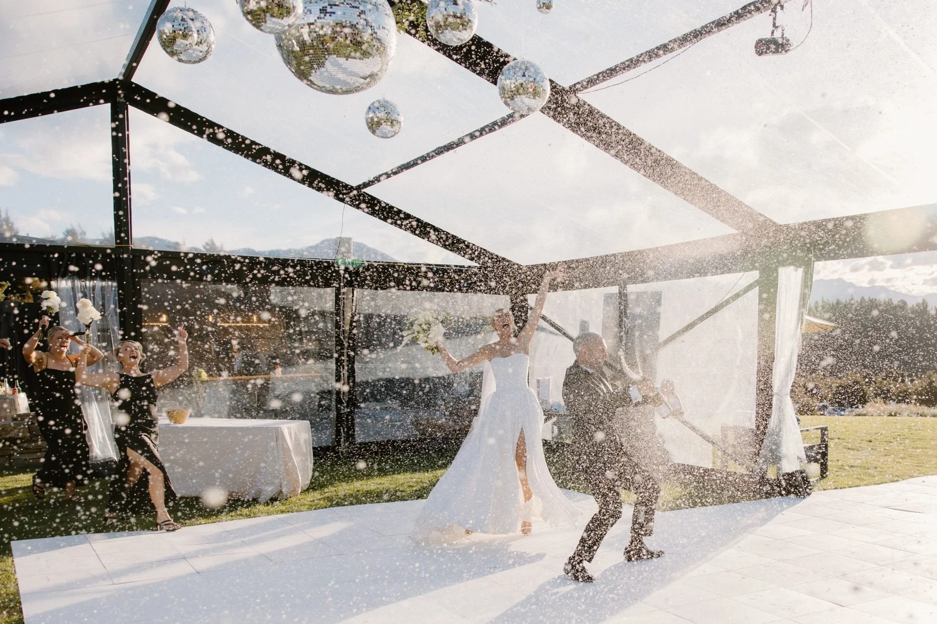 A bride and groom spray champagne as they enter their clear marquee reception at dublin bay in wanaka