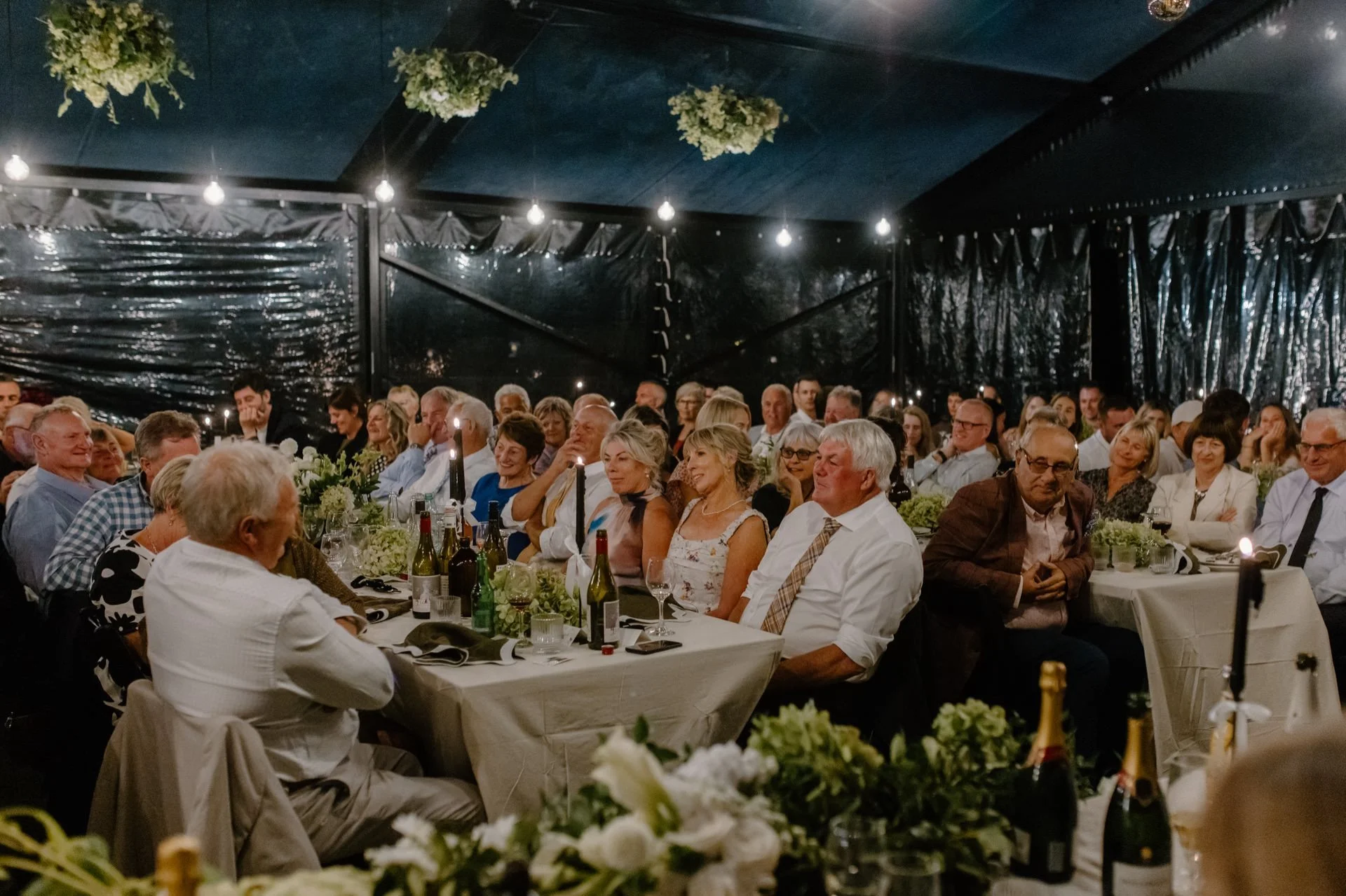 Guests sit listening to speeches under the cover of a clear marquee at dublin bay in wanaka for a summer wedding celebration.