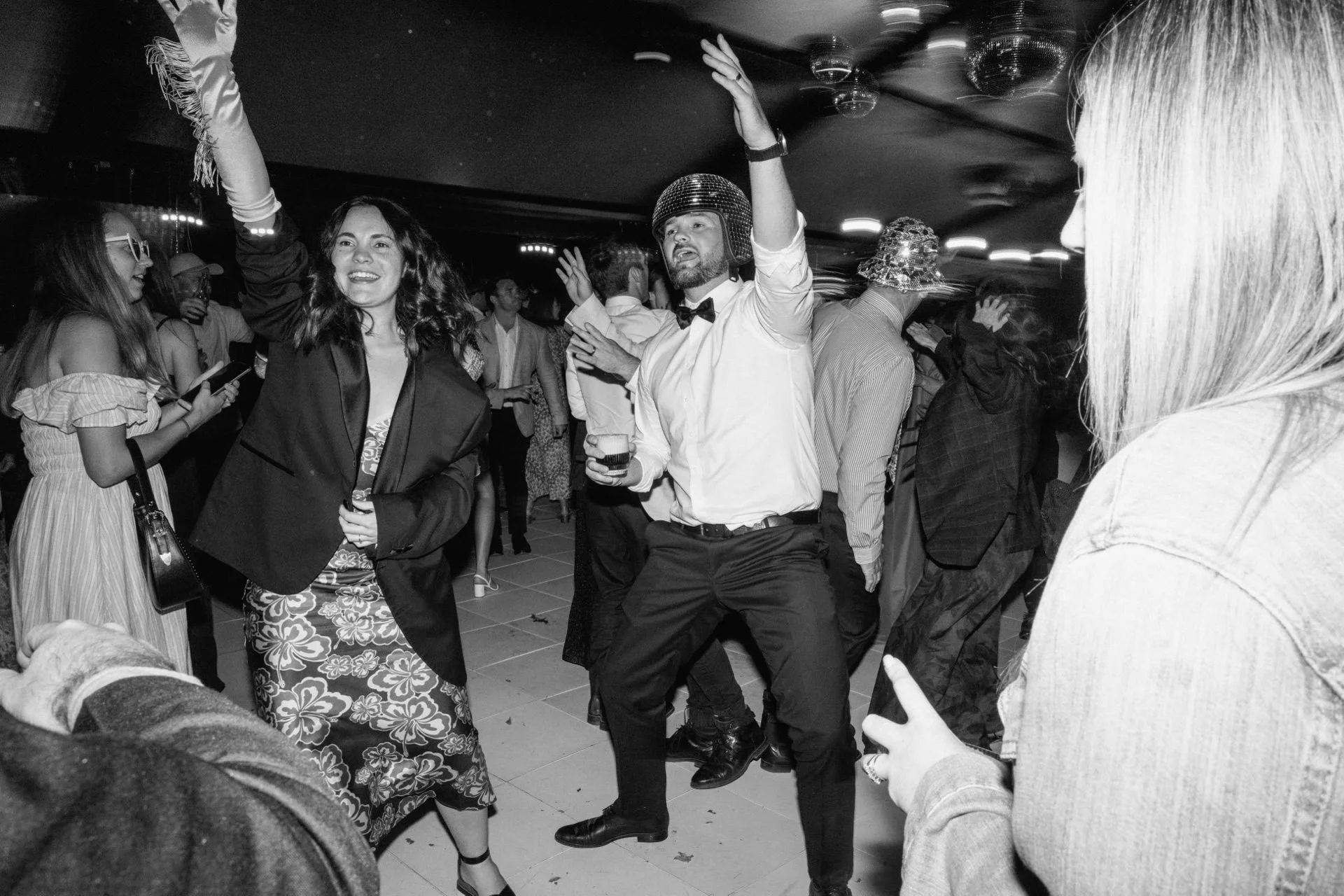 A black and white photo of a dance floor at a wedding at dublin bay wanaka. A guest is wearing a mirror ball helmet and other guests are dressed in fancy dress.
