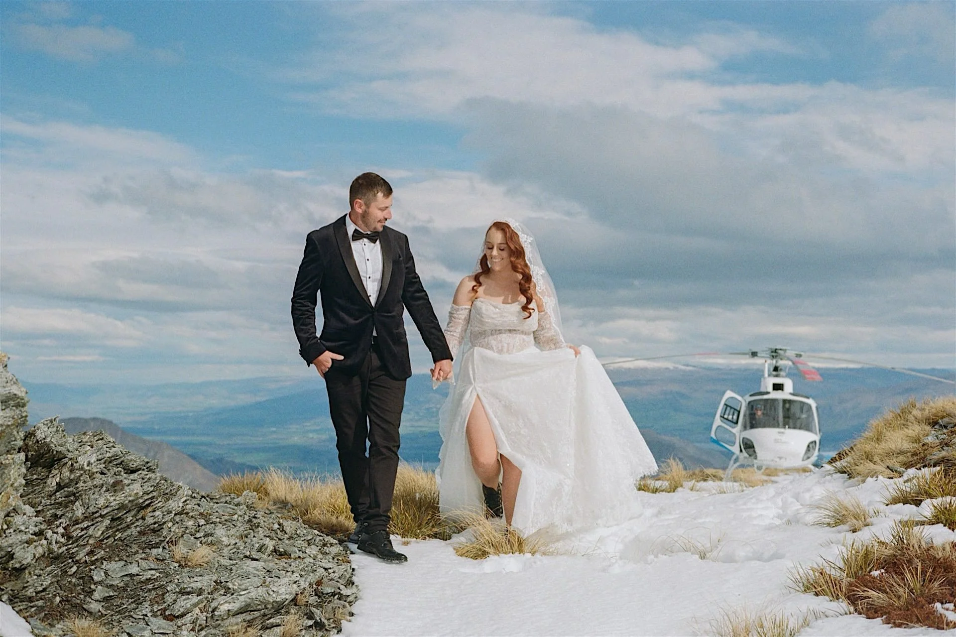  A married couple walk hand in hand on a snowy mountain in Wanaka with a helicopter behind them 