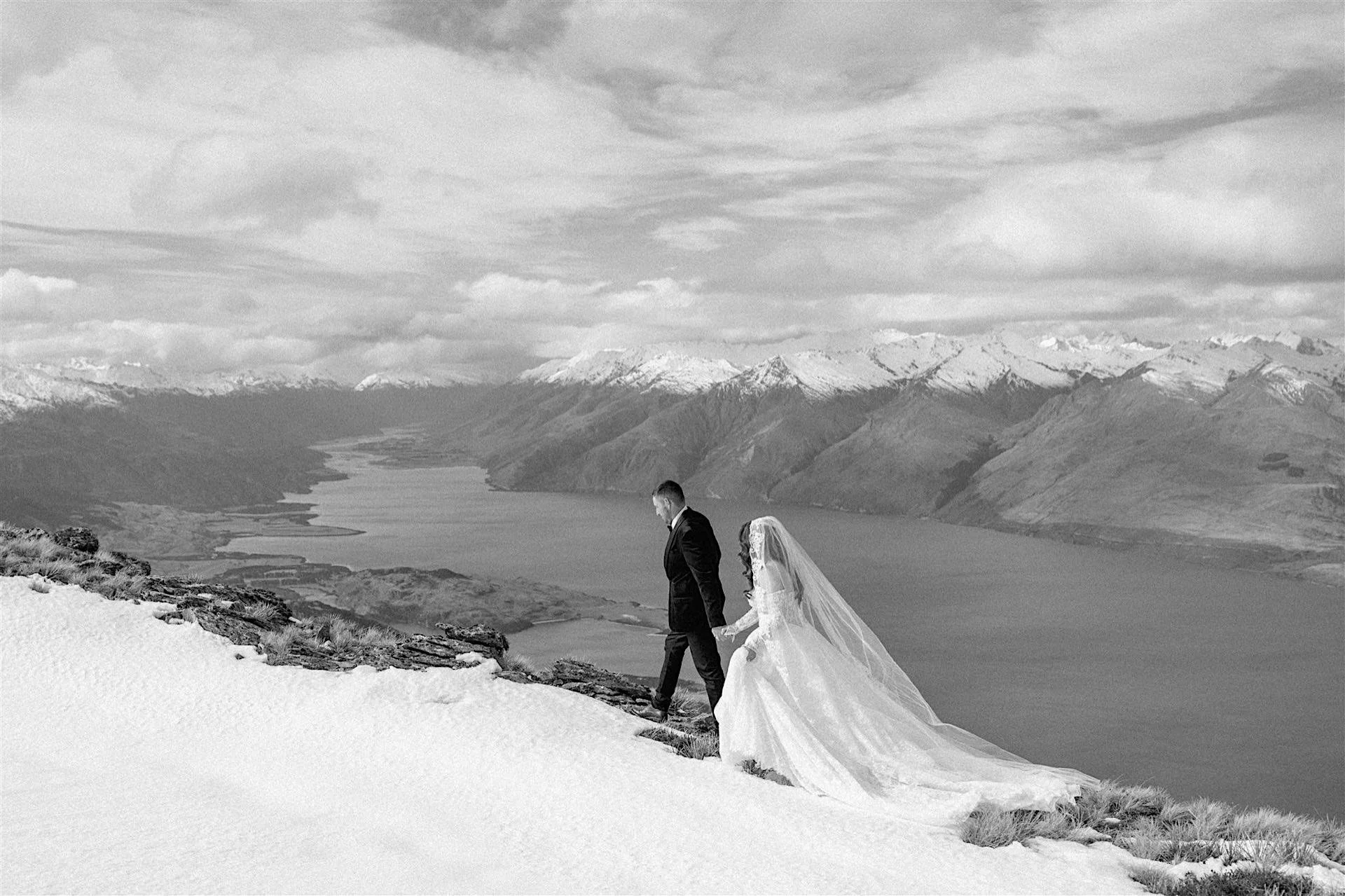  A black and white photo of a bride and groom on their wanaka wedding day walking along a ridgeline with snow on the mountains and lake wanaka behind them 