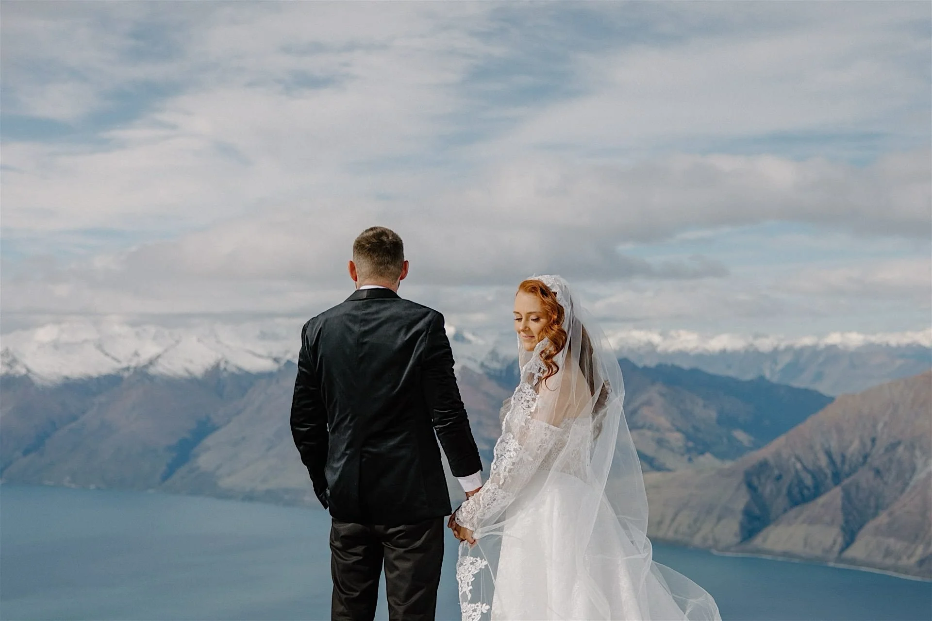  A bride looks over her shoulder as her groom admires the view high on a snowy mountain in Wanaka, New zealand on their wedding day 