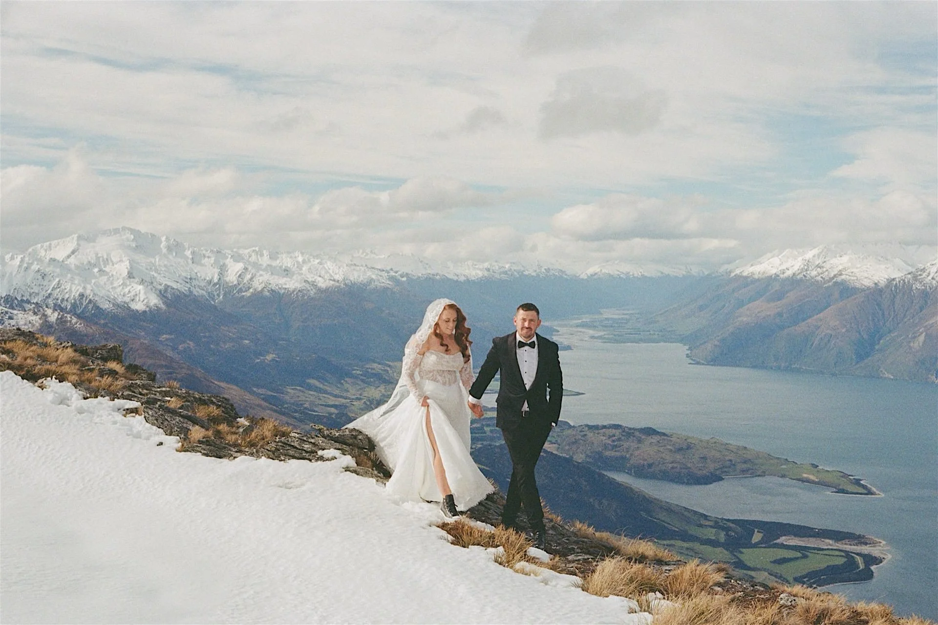  A 35mm film photo of a newly married couple on their wanaka elopement.  They walk hand in hand along a snowy ridgeline with lake wanaka behind them 