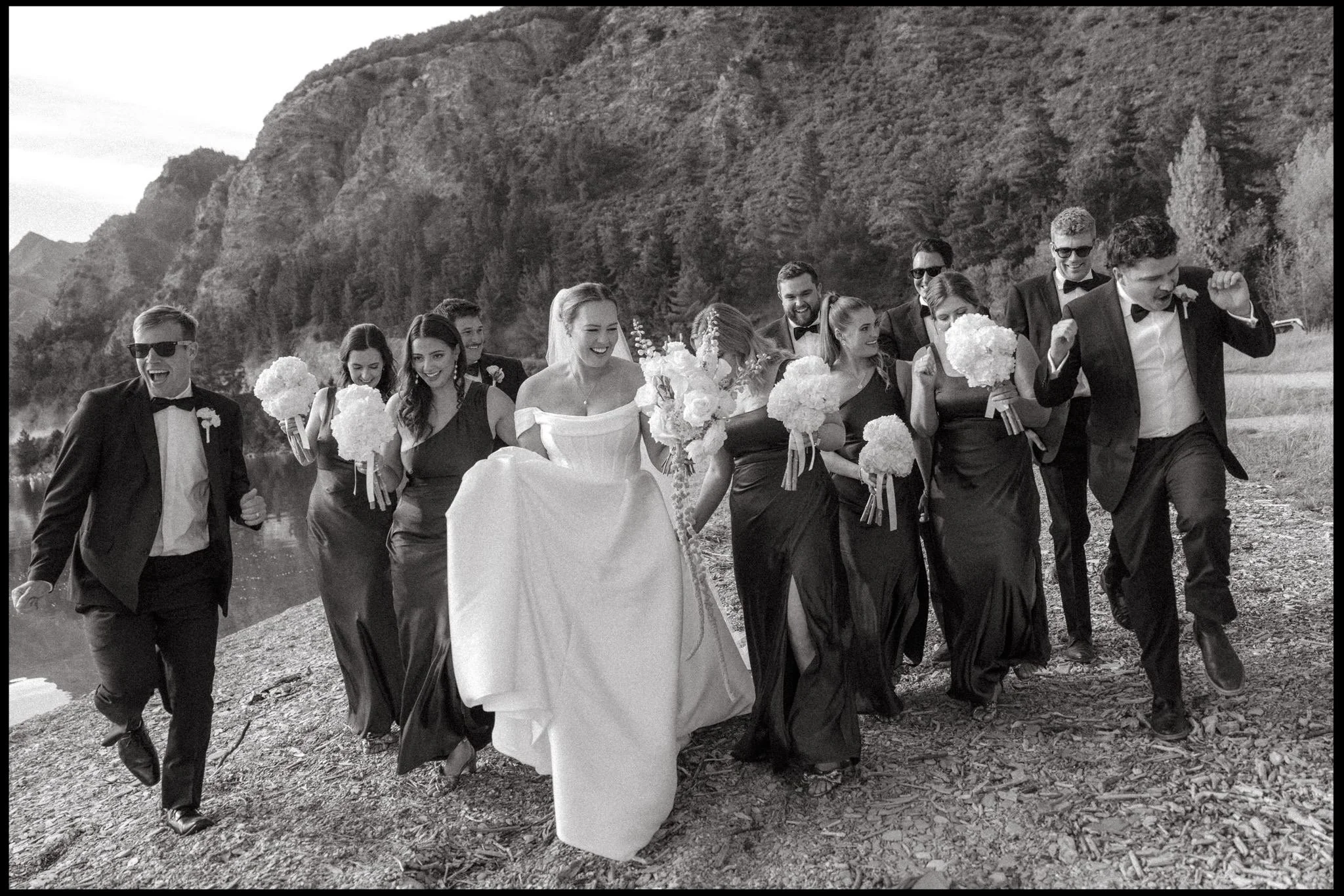 A bride and groom walk with their bridal party along the shore of lake Hawea on their autumn wedding day