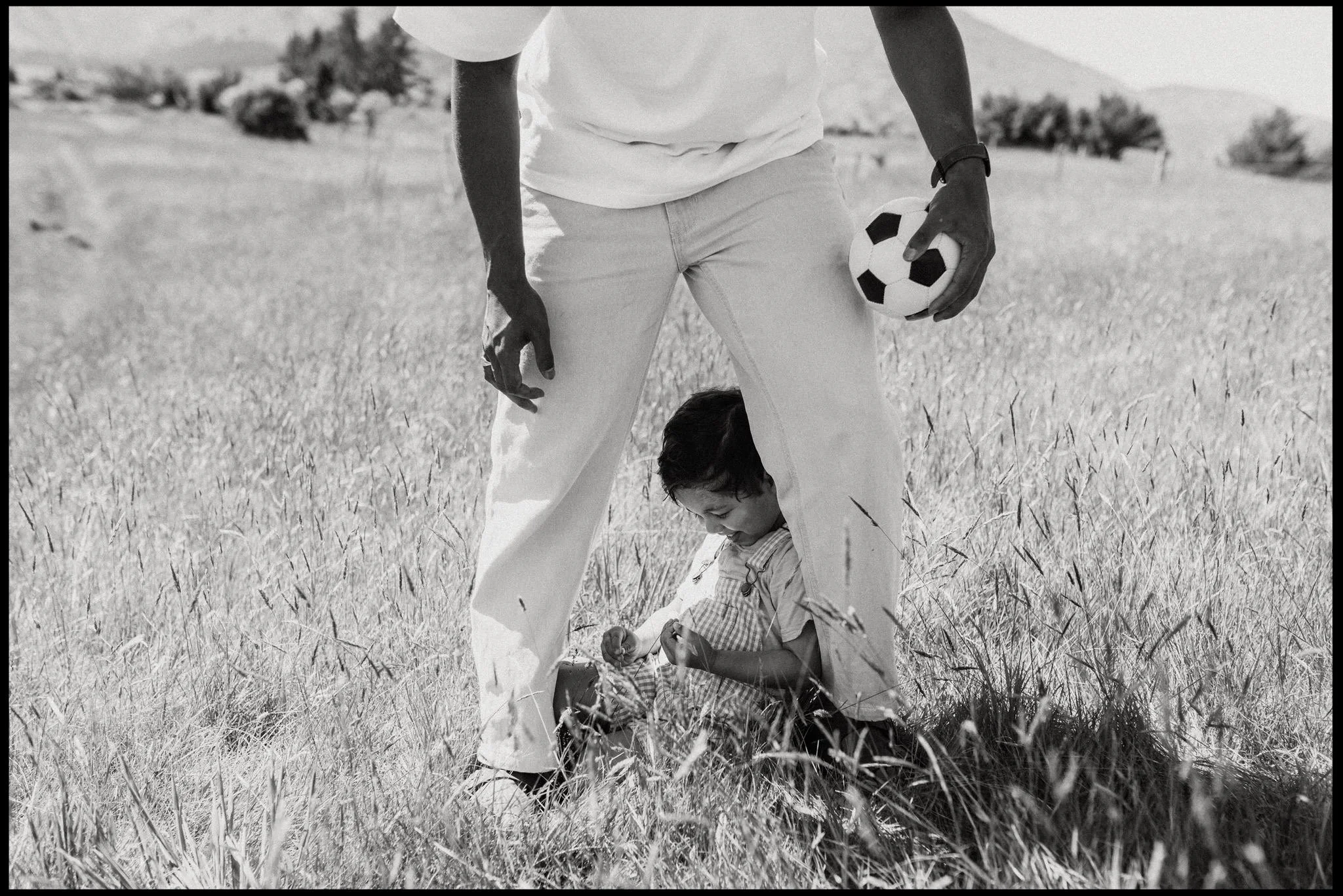 on a family portrait session in Wanaka a little boy sits between his dads legs in long grass on a wanaka summers day