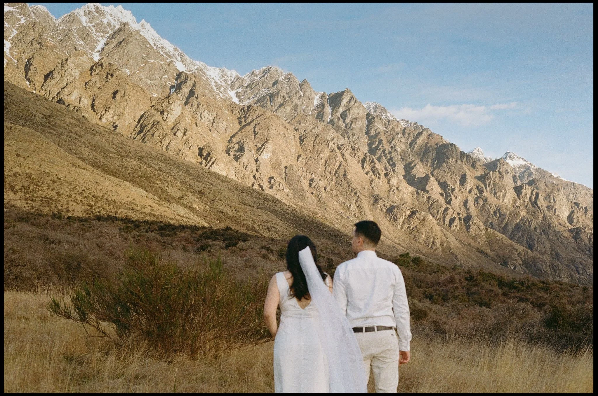 A bride and groom walk toward the towering mountains of queenstown on their wedding day at sunset