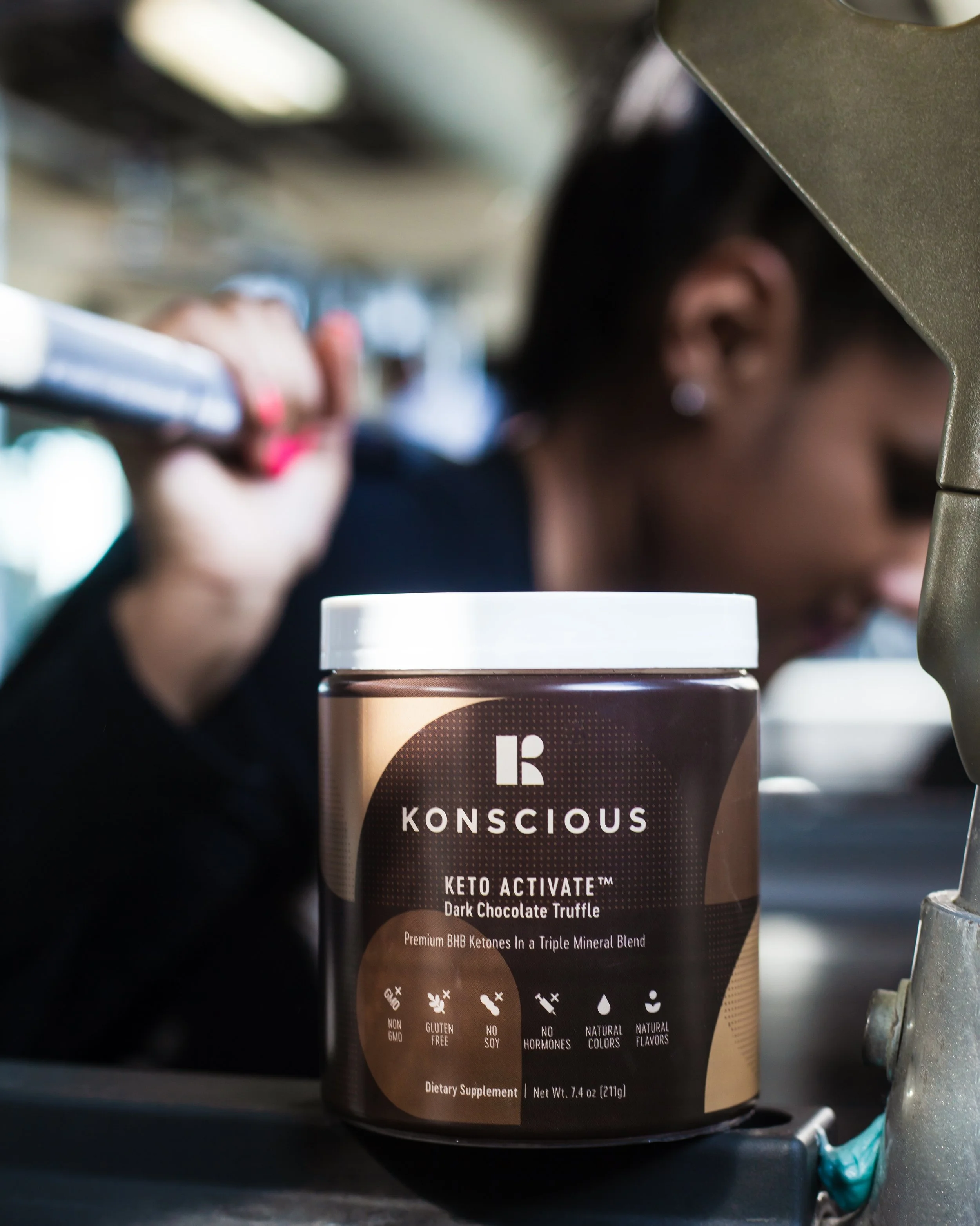 A container of KONSCIOUS Keto Activate Dark Chocolate Truffle dietary supplement in front of a woman with dark hair and earrings, lifting a barbell in a gym.
