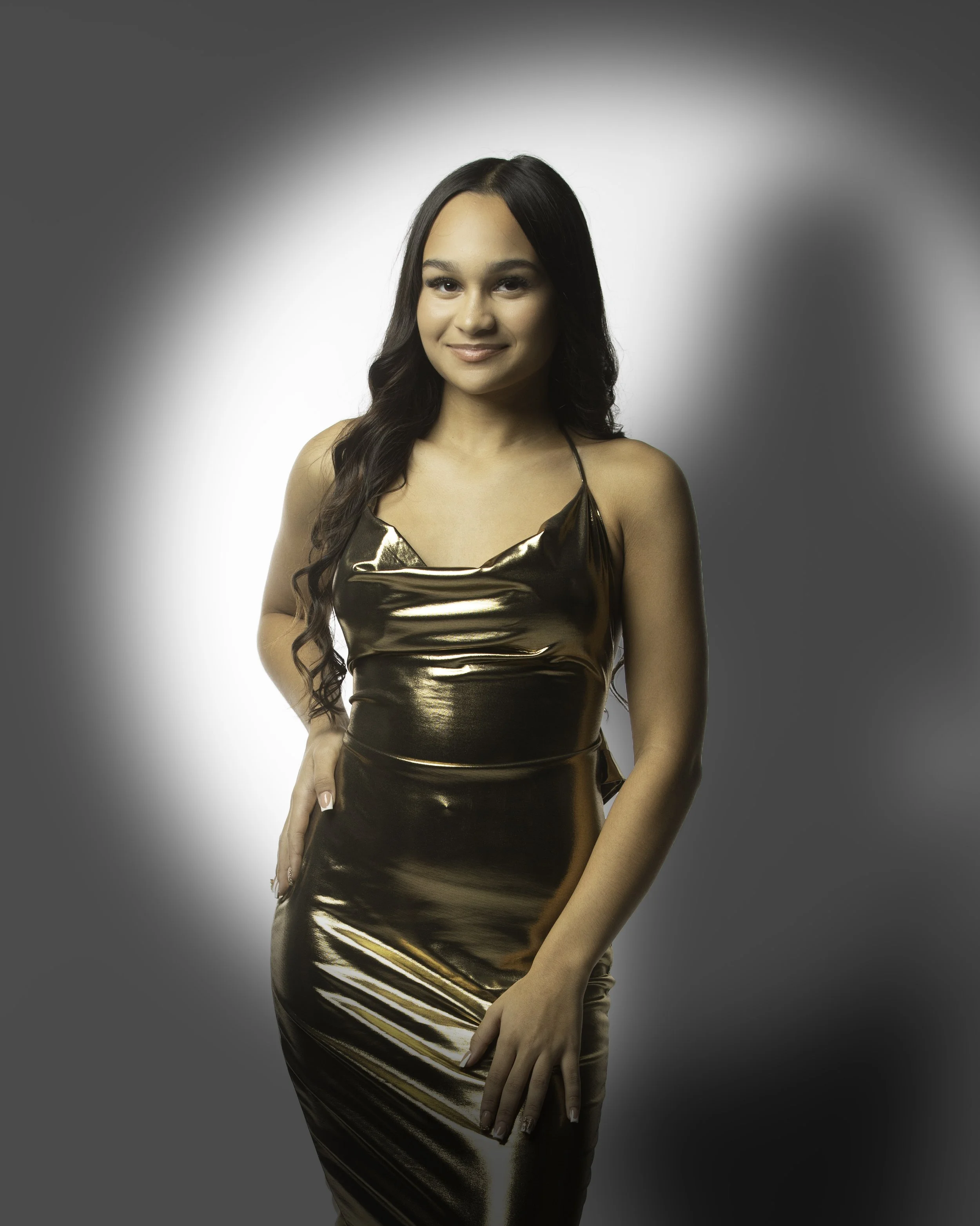 A young woman with long dark hair in a black and gold shiny dress posing against a gray background.