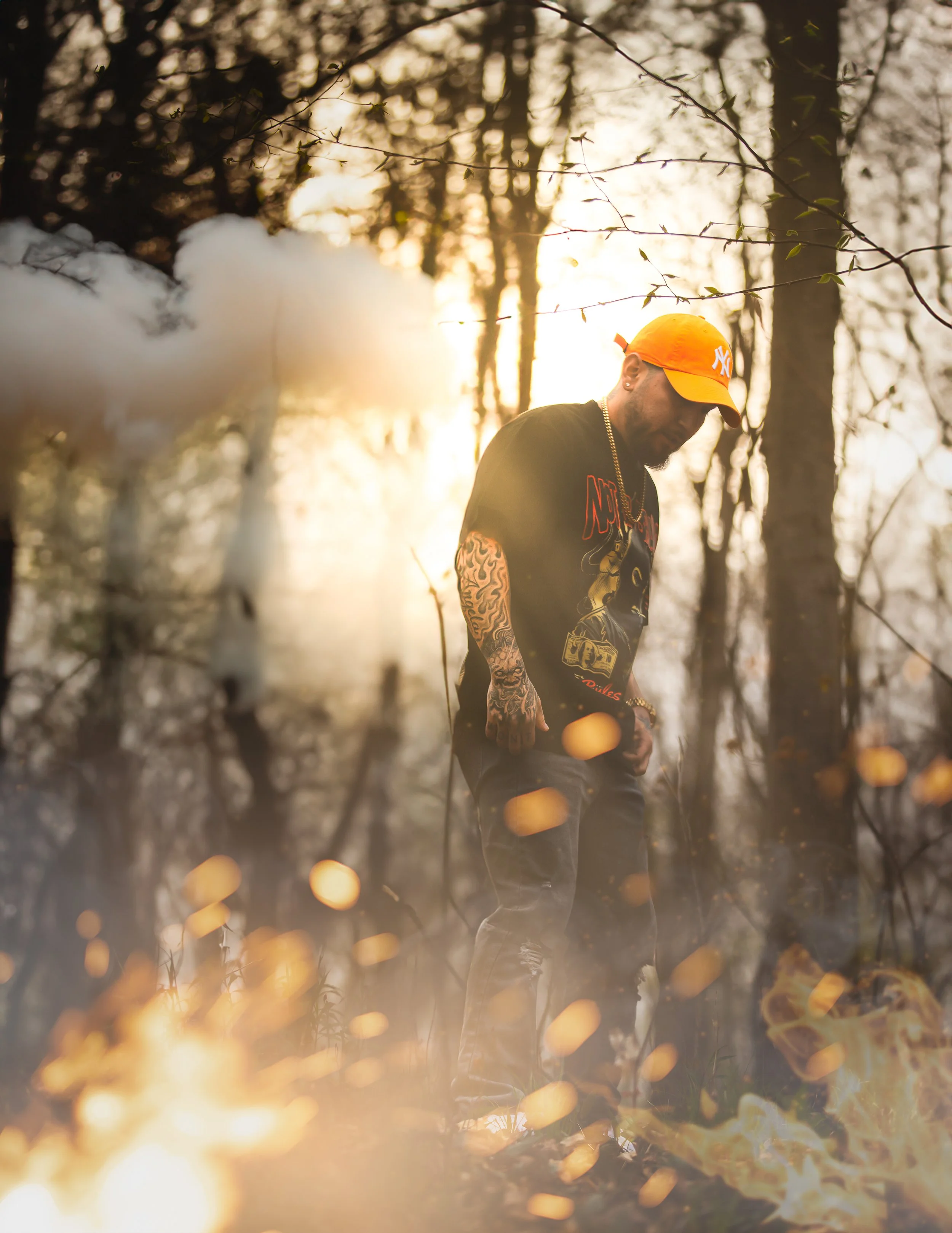 A man with tattoos on his arm, wearing a black graphic t-shirt, ripped jeans, an orange New York Yankees cap, and gold jewelry, stands in a wooded area during sunset. Smoke and fire are visible around him.