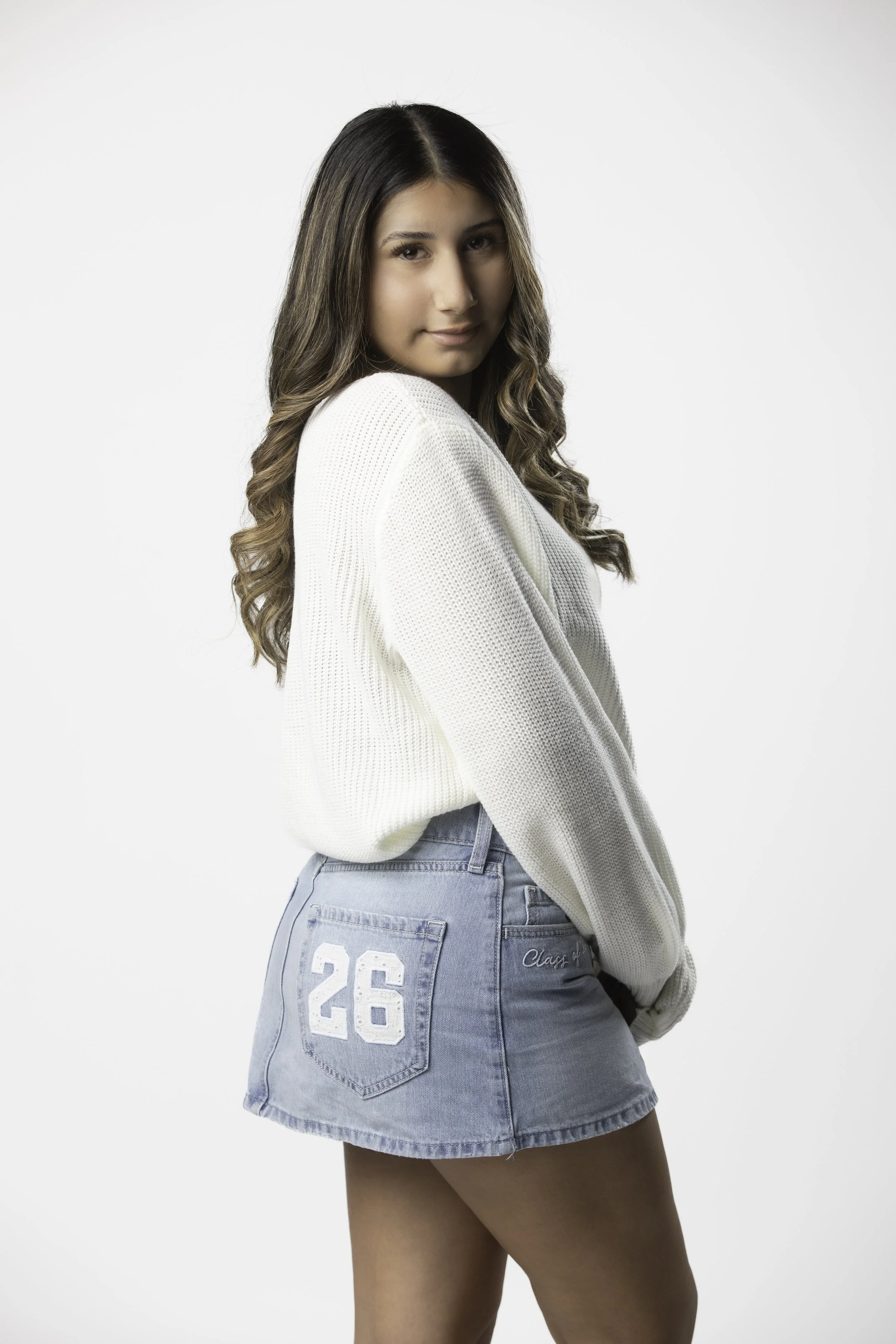 A young woman with long, wavy brown hair wearing a white sweater and a denim skirt with the number 26 on the back, standing against a plain white background.