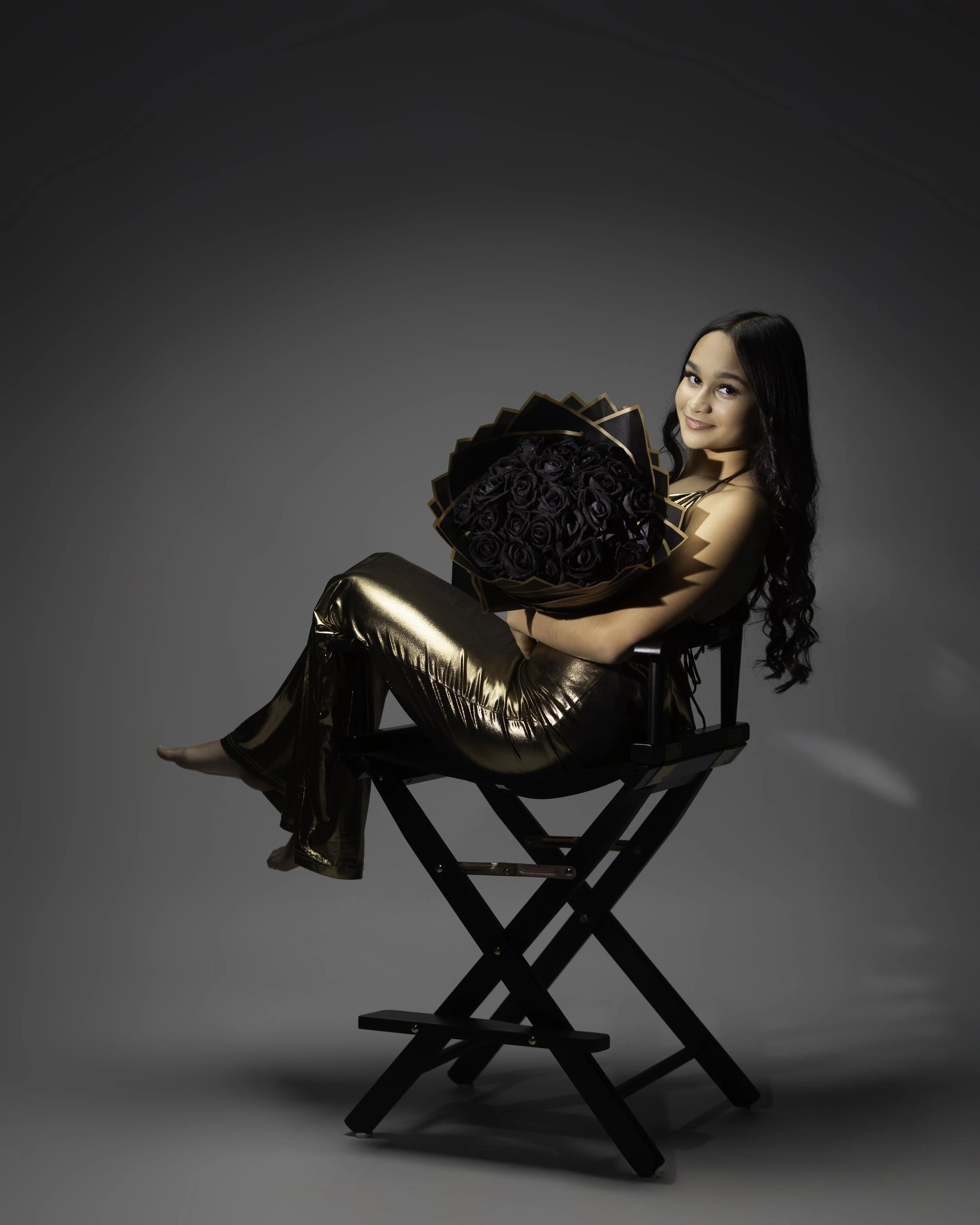 A woman sitting on a black director's chair, holding a bouquet of black roses, dressed in shiny gold pants, with long dark curly hair and a slight smile, against a plain grey background.