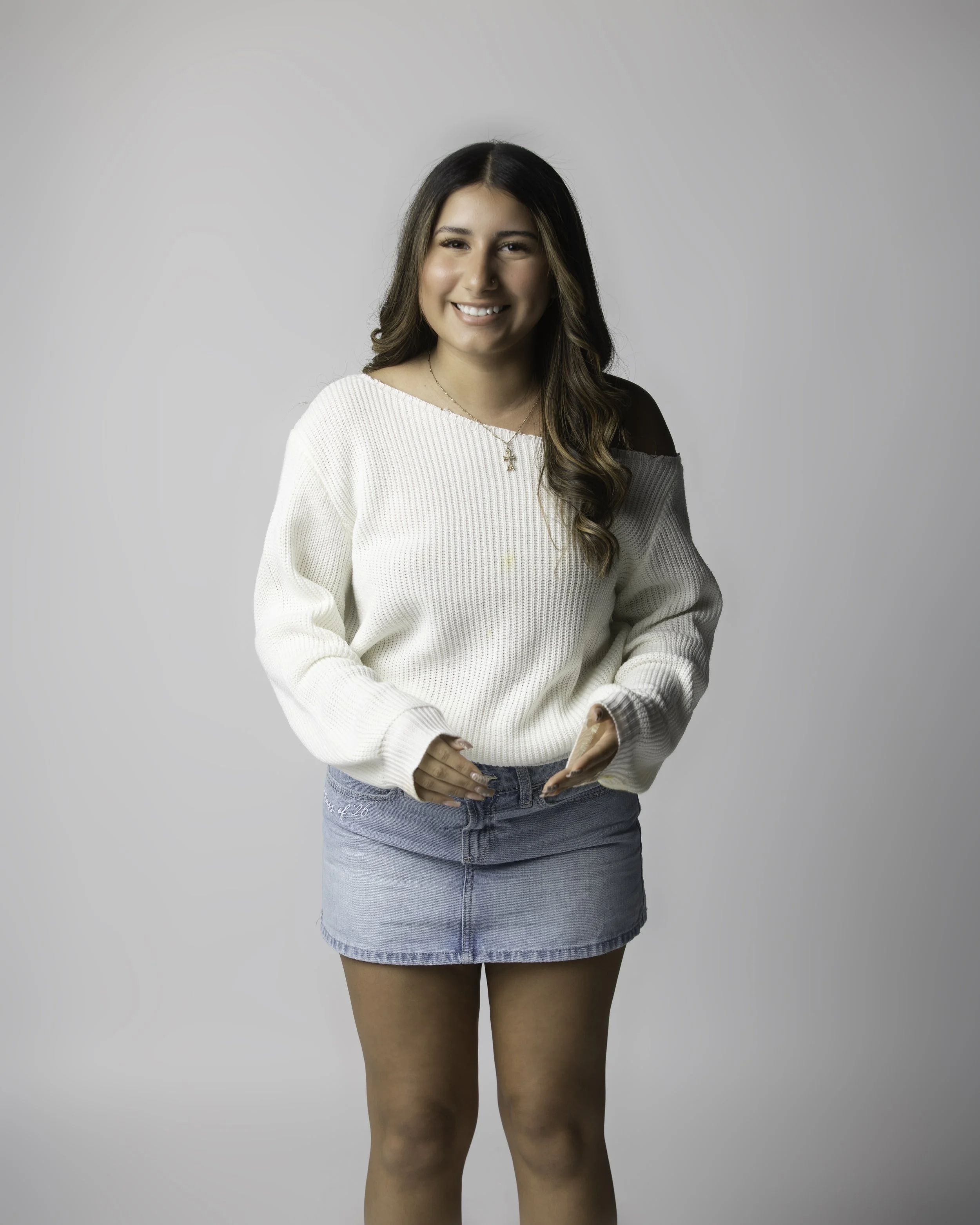 A young woman with long wavy brown hair, smiling, wearing a white sweater with cold shoulder design and a light blue denim skirt, standing against a plain neutral background.