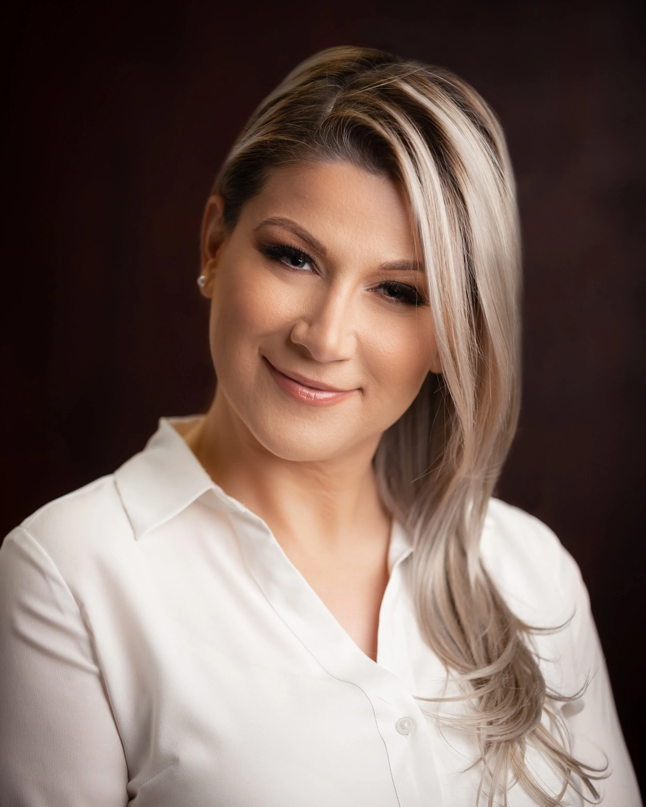 A woman with blonde hair styled in loose waves, wearing a white blouse, against a dark background.